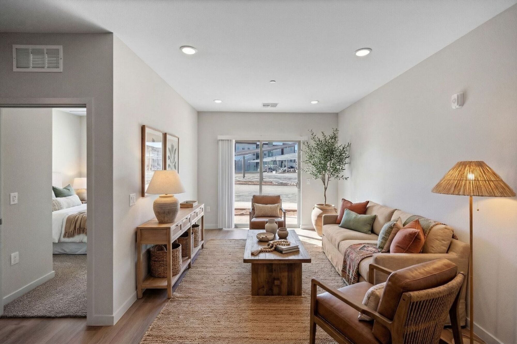 Bright living room with neutral decor, wooden furniture, and large window beside a bedroom entry.