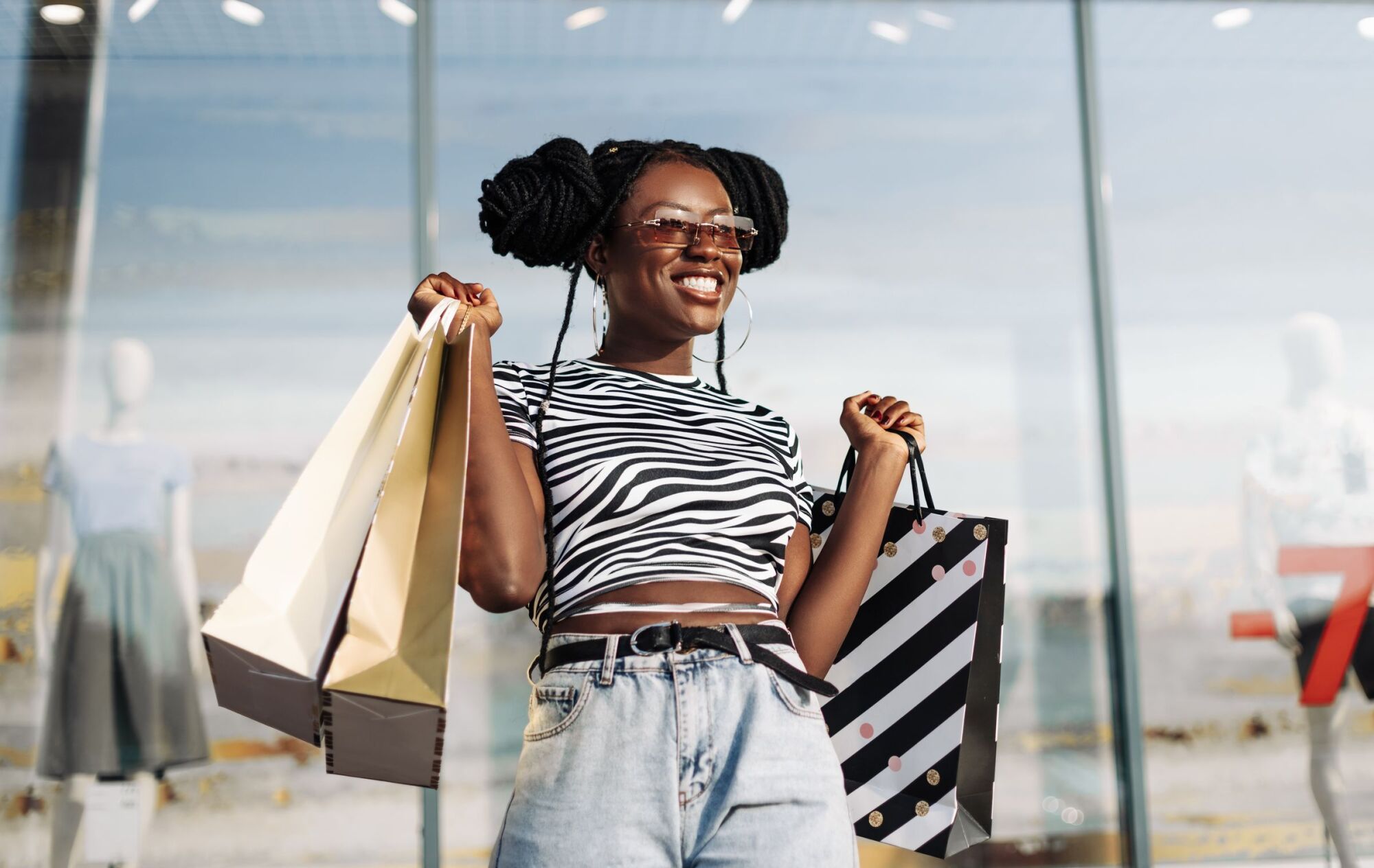 Smiling woman in zebra print top holds shopping bags outside a store, wearing sunglasses and jeans.
