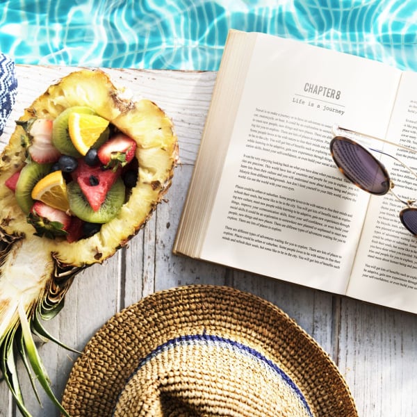 A pineapple is cut open and filled with fruit and placed next to a book and sunglasses.