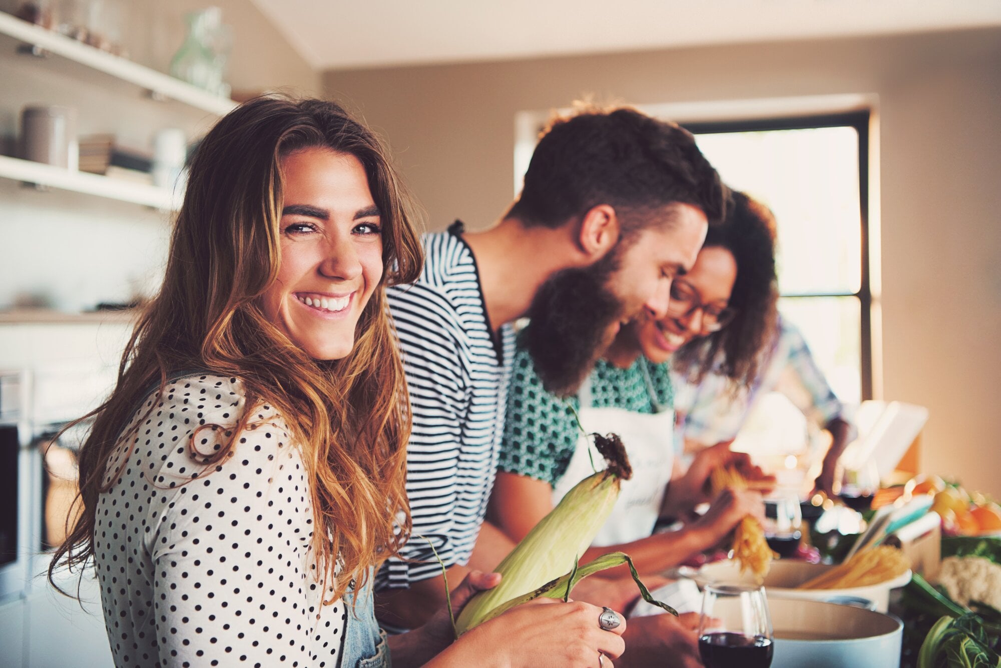A woman with a polka dot shirt is holding a corn cob while two people prepare food in the background.