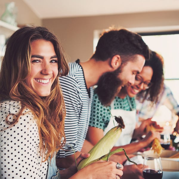 A woman with a polka dot shirt is holding a corn cob while two people prepare food in the background.