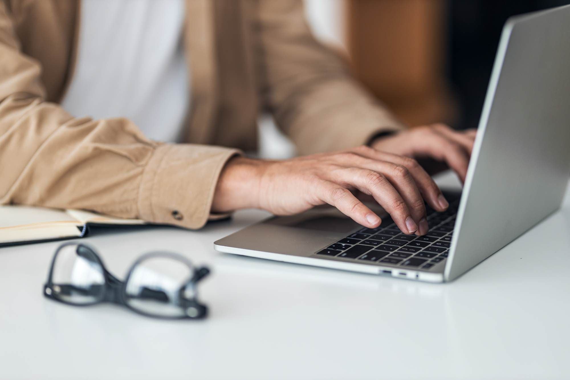 Person typing on a laptop at a desk with glasses and a notebook nearby
