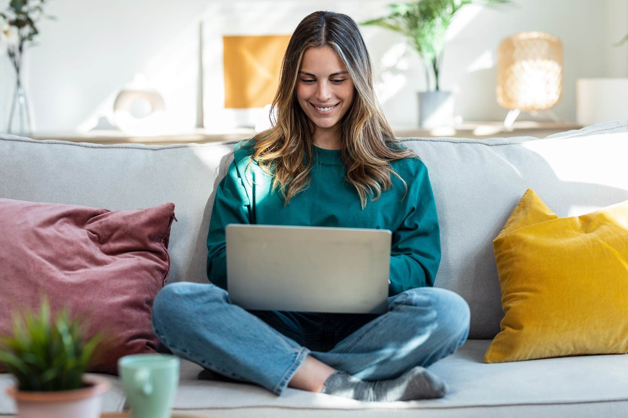 Woman sitting cross-legged on a sofa, smiling while using a laptop in a bright living room