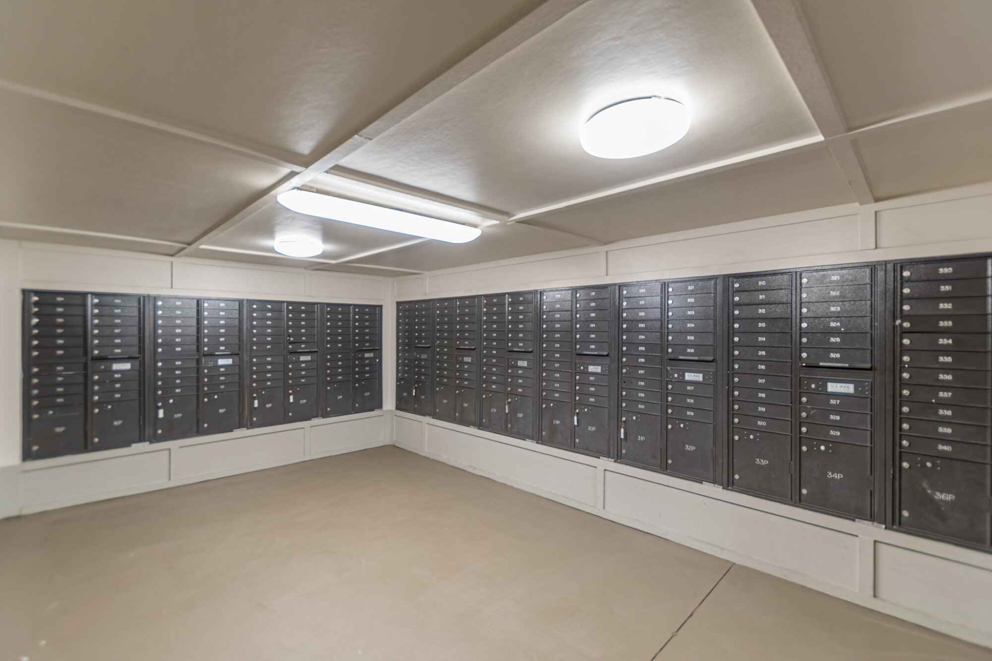 A corner package room with rows of black metal mailboxes lining two white walls under bright ceiling lights at Apex West Midtown.