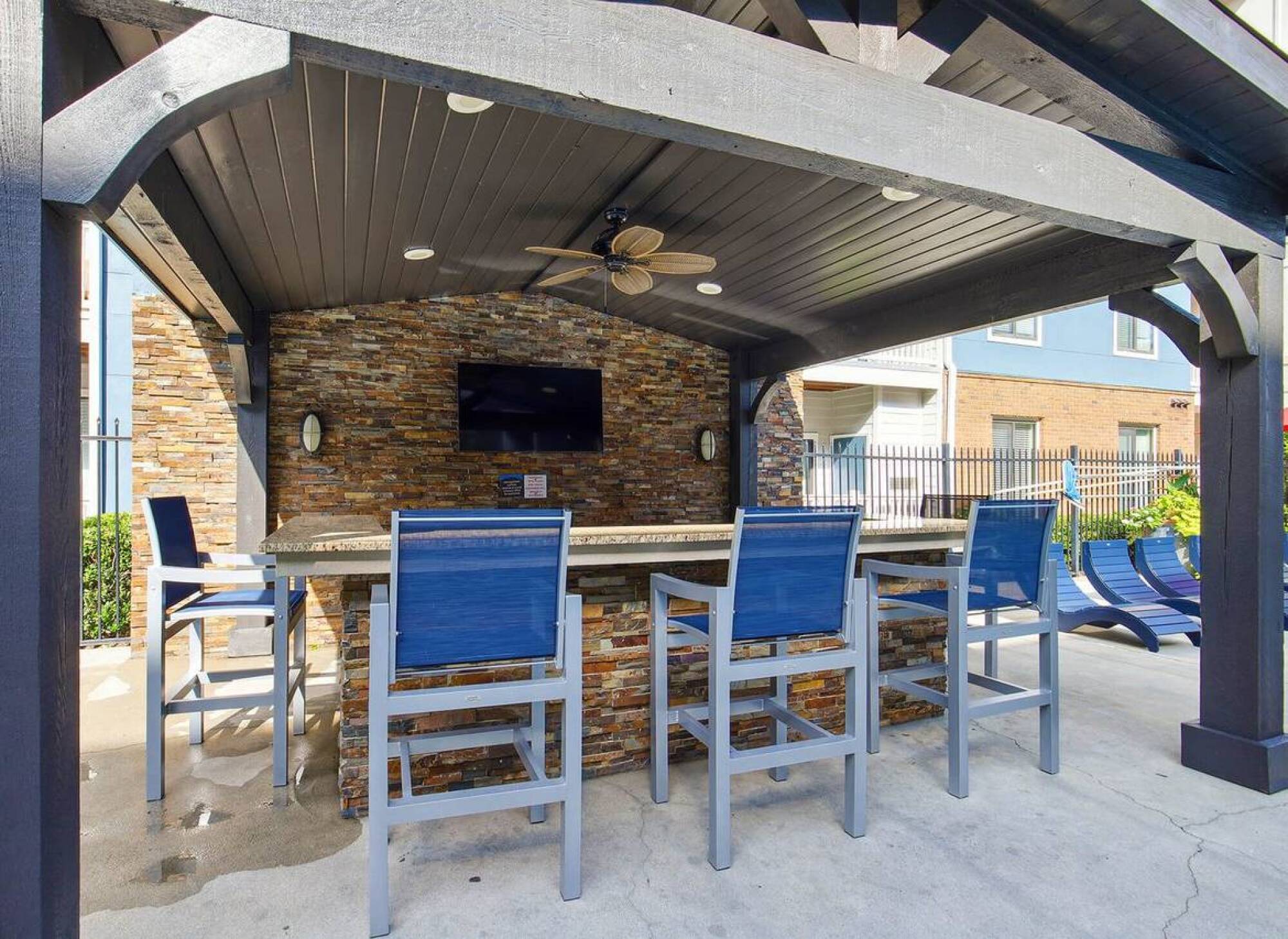 Apex West Midtown outdoor bar area with stone counter, blue chairs, TV, ceiling fan, and shaded pergola.