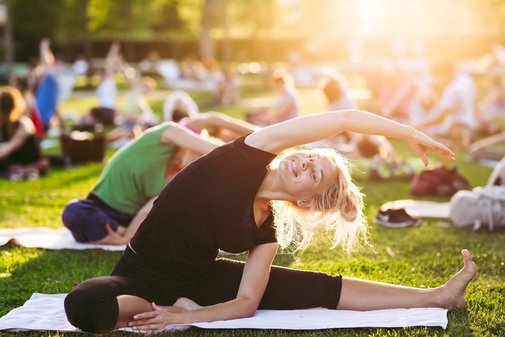 Woman stretching on a yoga mat outdoors in a park with others, during a sunny group yoga session.