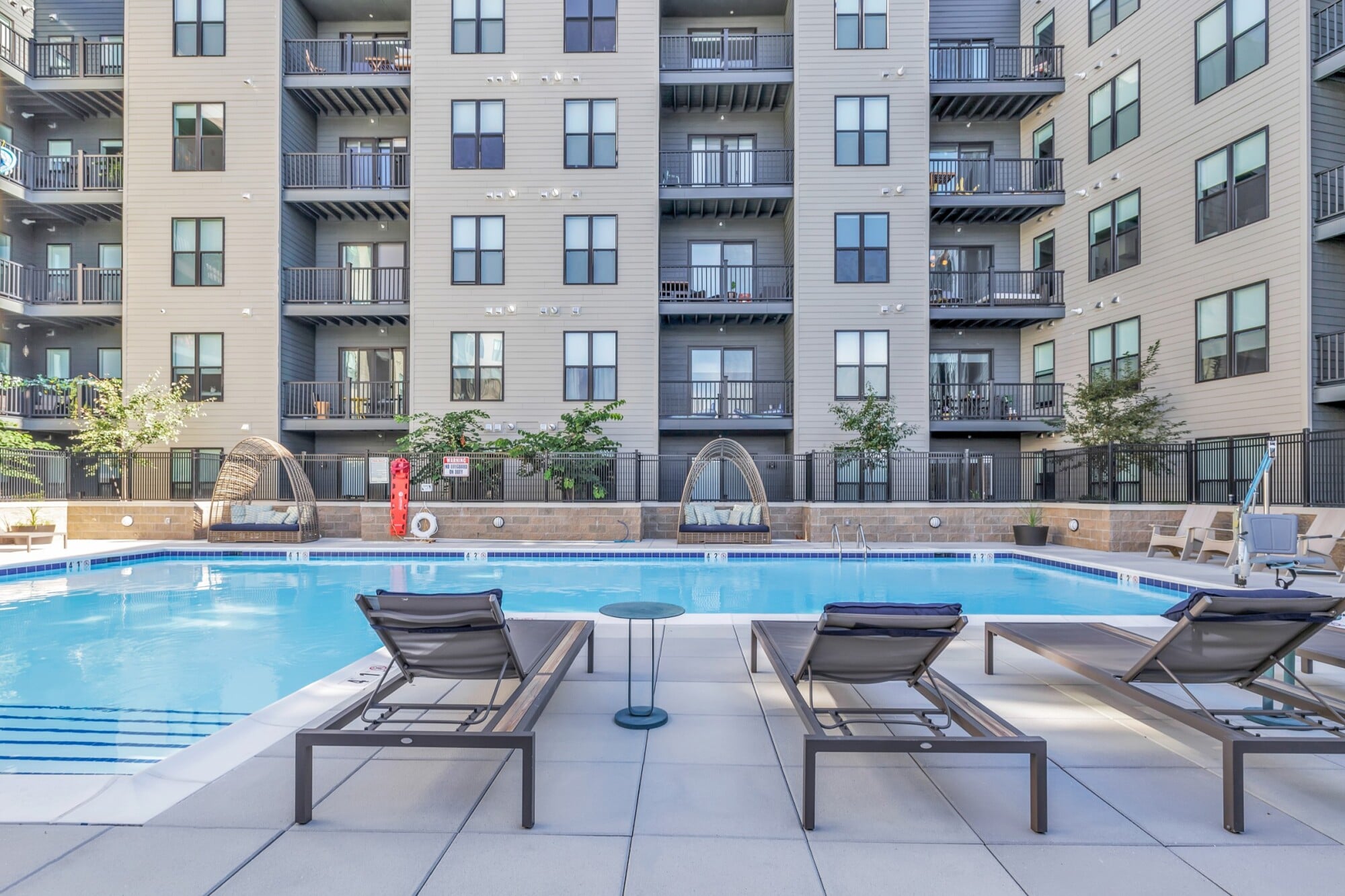 Modern apartment building with balconies, pool, lounge chairs, and cabanas on a sunny day.