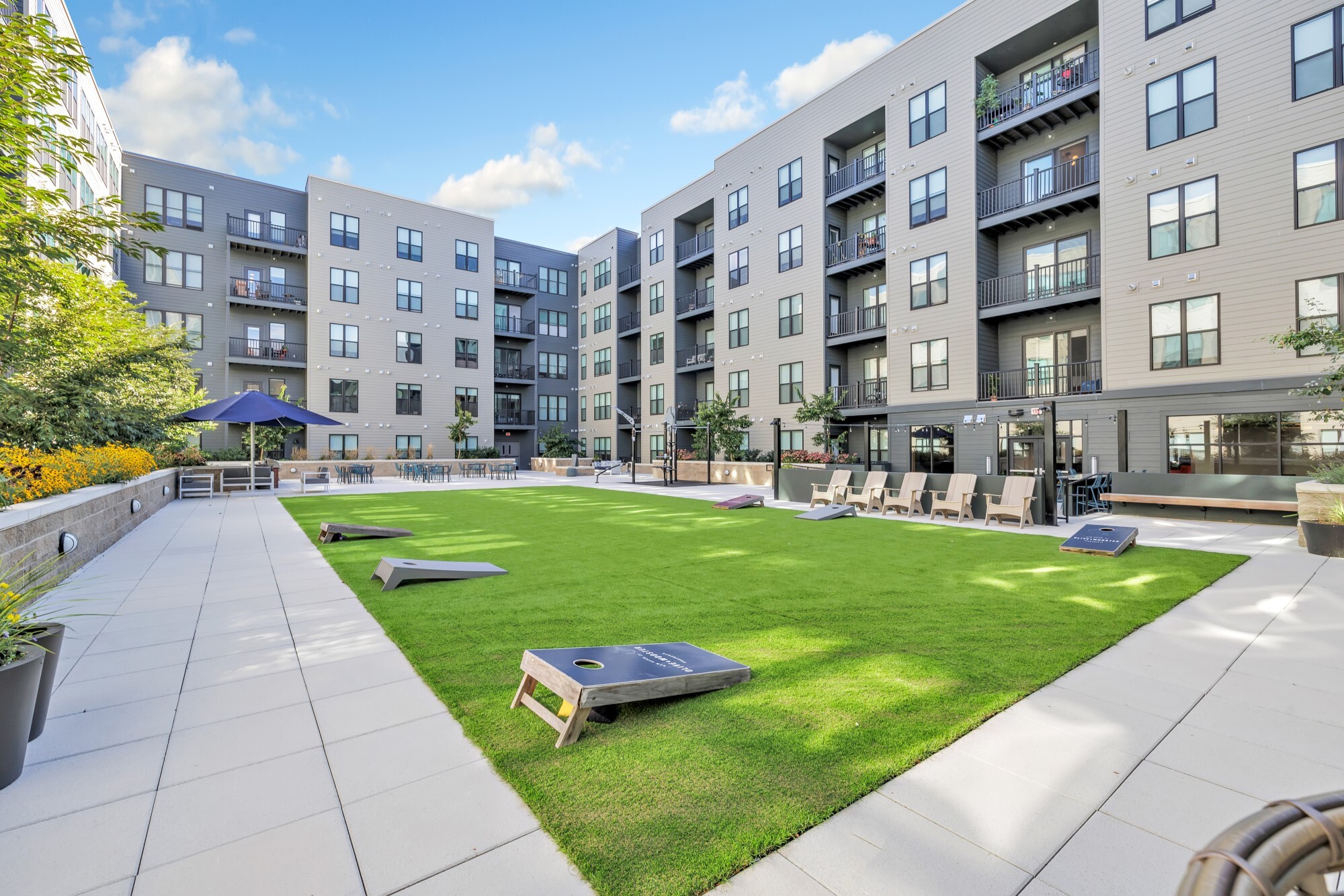 Outdoor courtyard with green lawn, cornhole boards, lounge chairs, and modern apartment buildings surrounding the area.