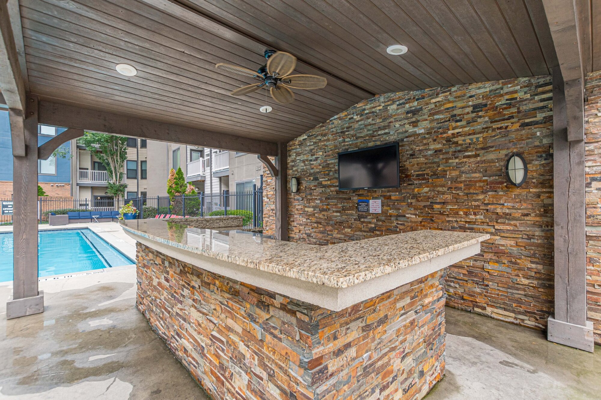Outdoor stone bar with granite countertop under a wooden pergola, next to a swimming pool and TV on the wall at Apex West Midtown.