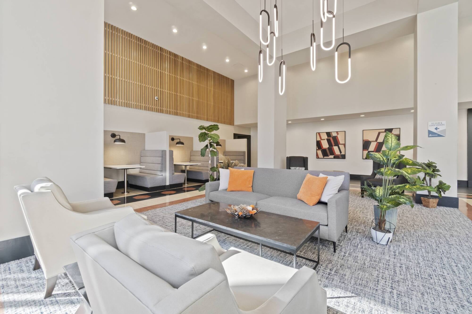 Modern lobby with gray sofas, orange pillows, white chairs, plants, artwork, and pendant lights in a bright, airy space.