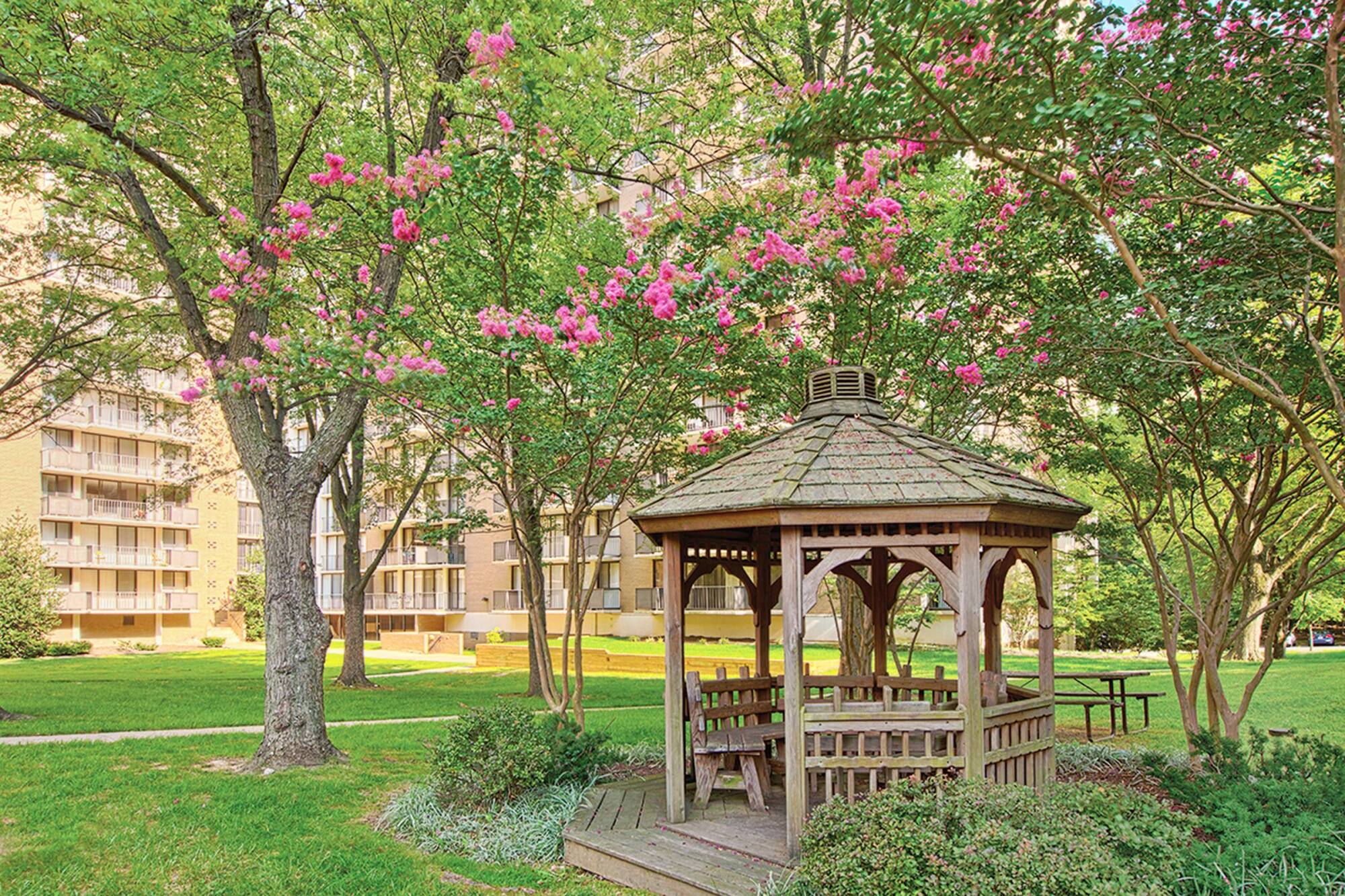 Milestone Apartments White gazebo entrance surrounded by greenery, leading to tennis courts on a sunny day.