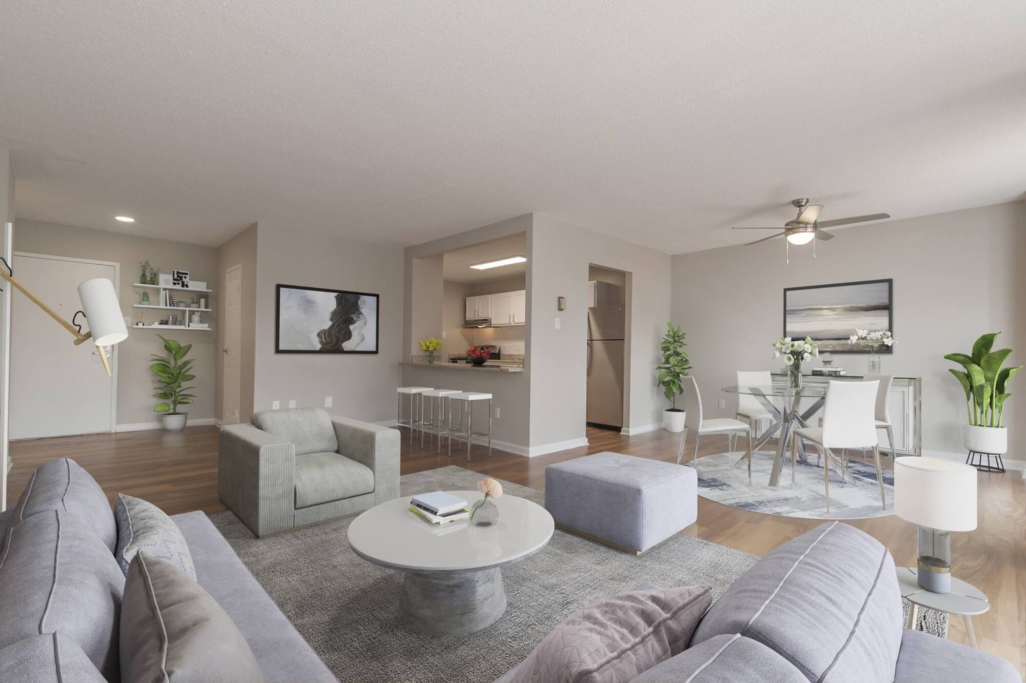 Modern living room with gray furniture, an open kitchen, dining area, plants, and large windows letting in natural light.