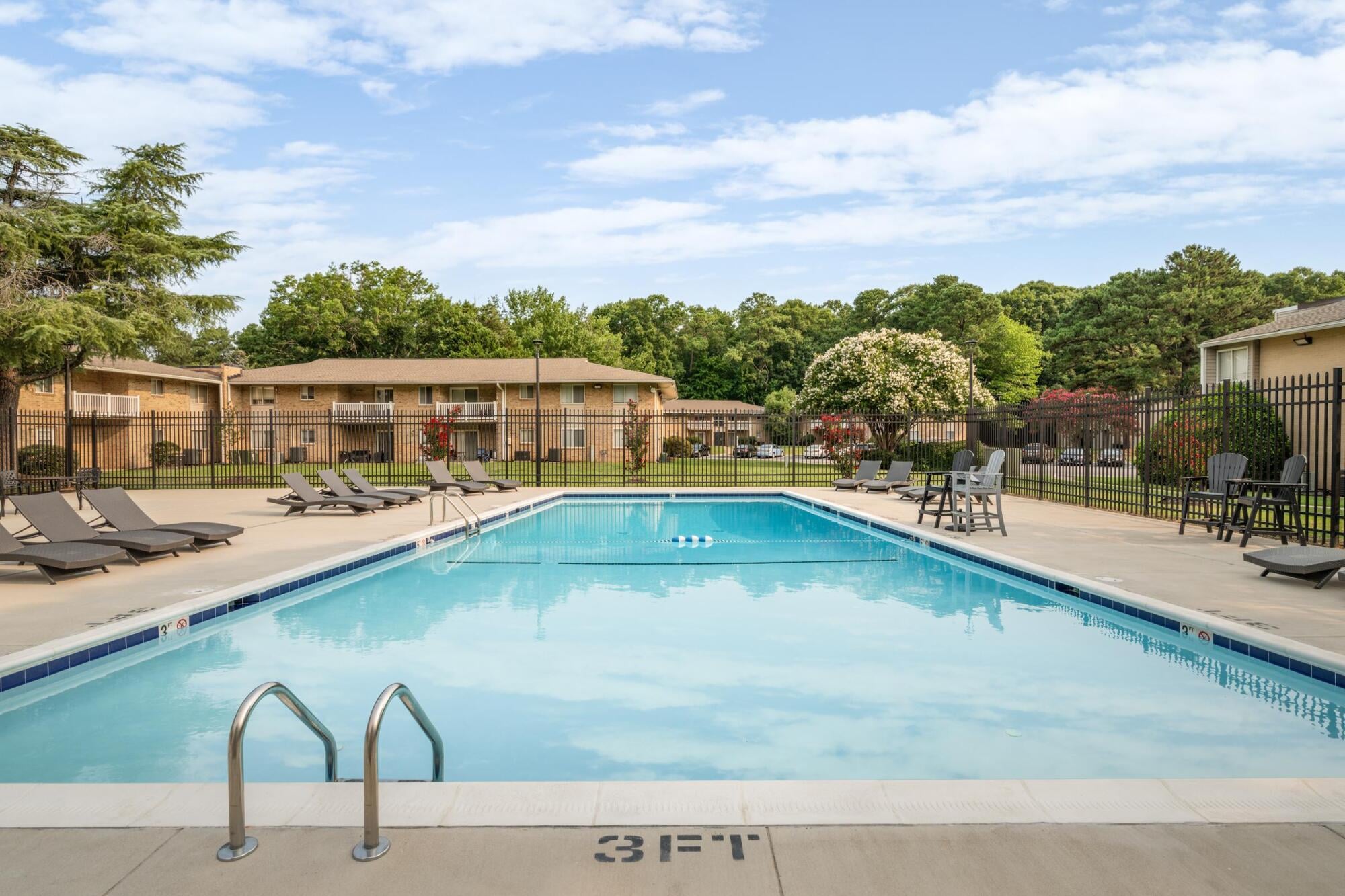 Outdoor pool with lounge chairs and apartments in the background, surrounded by trees and a fenced area, under a partly cloudy sky.