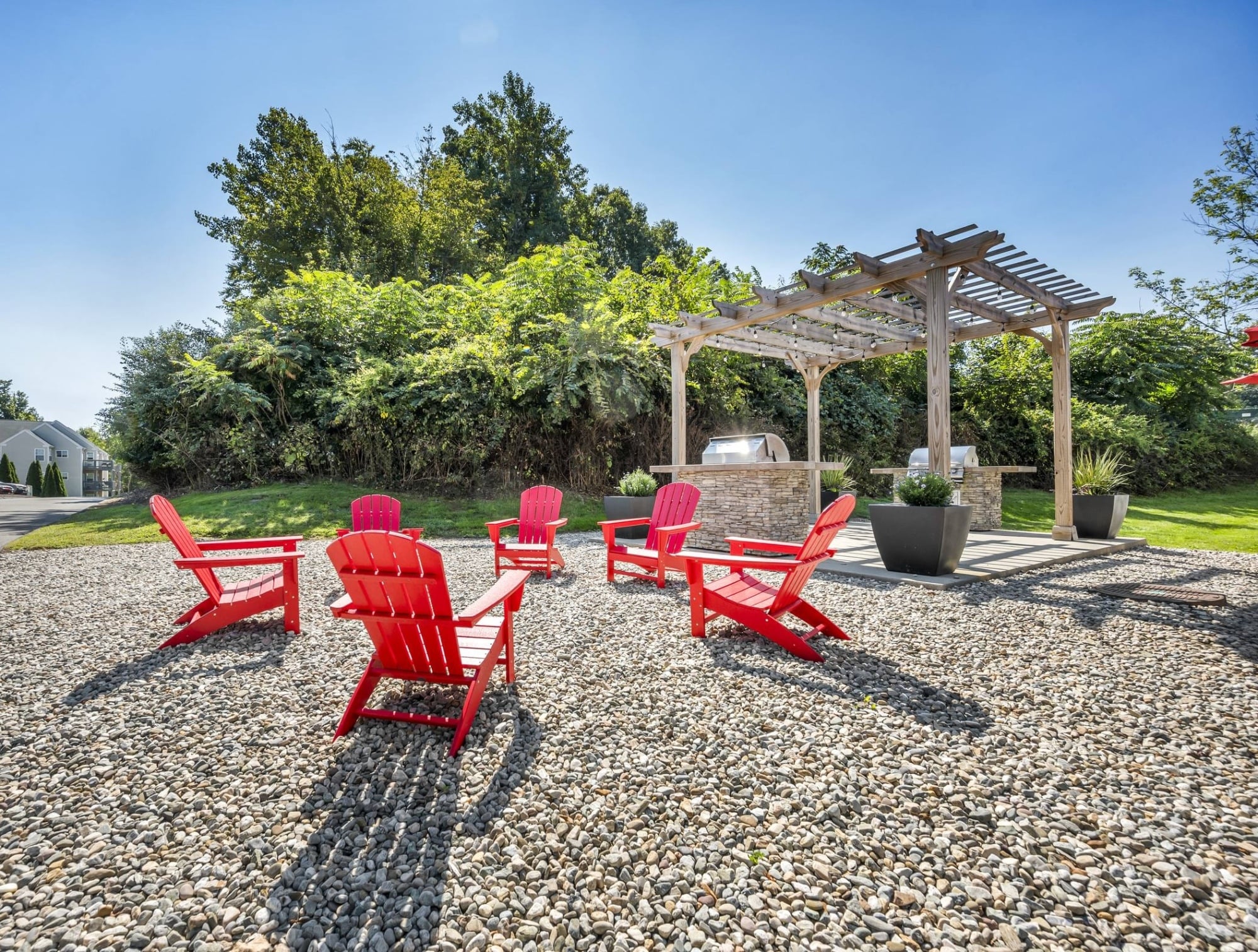 Six red Adirondack chairs on gravel, arranged around a fire pit, with a pergola and barbecue grill in the background.