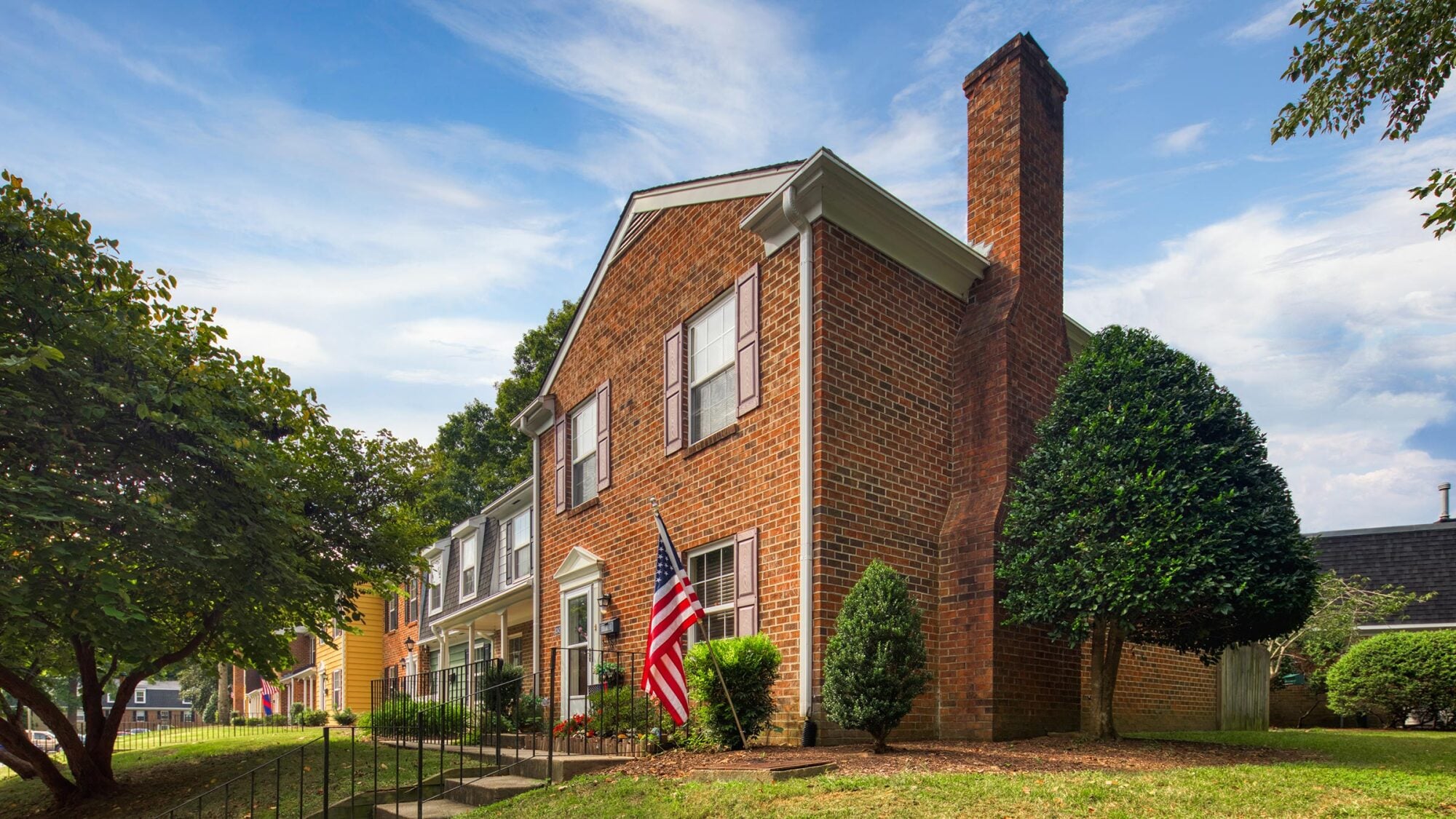 Two-story brick house with a large chimney, American flag, front yard, and shrubs under a partly cloudy sky.