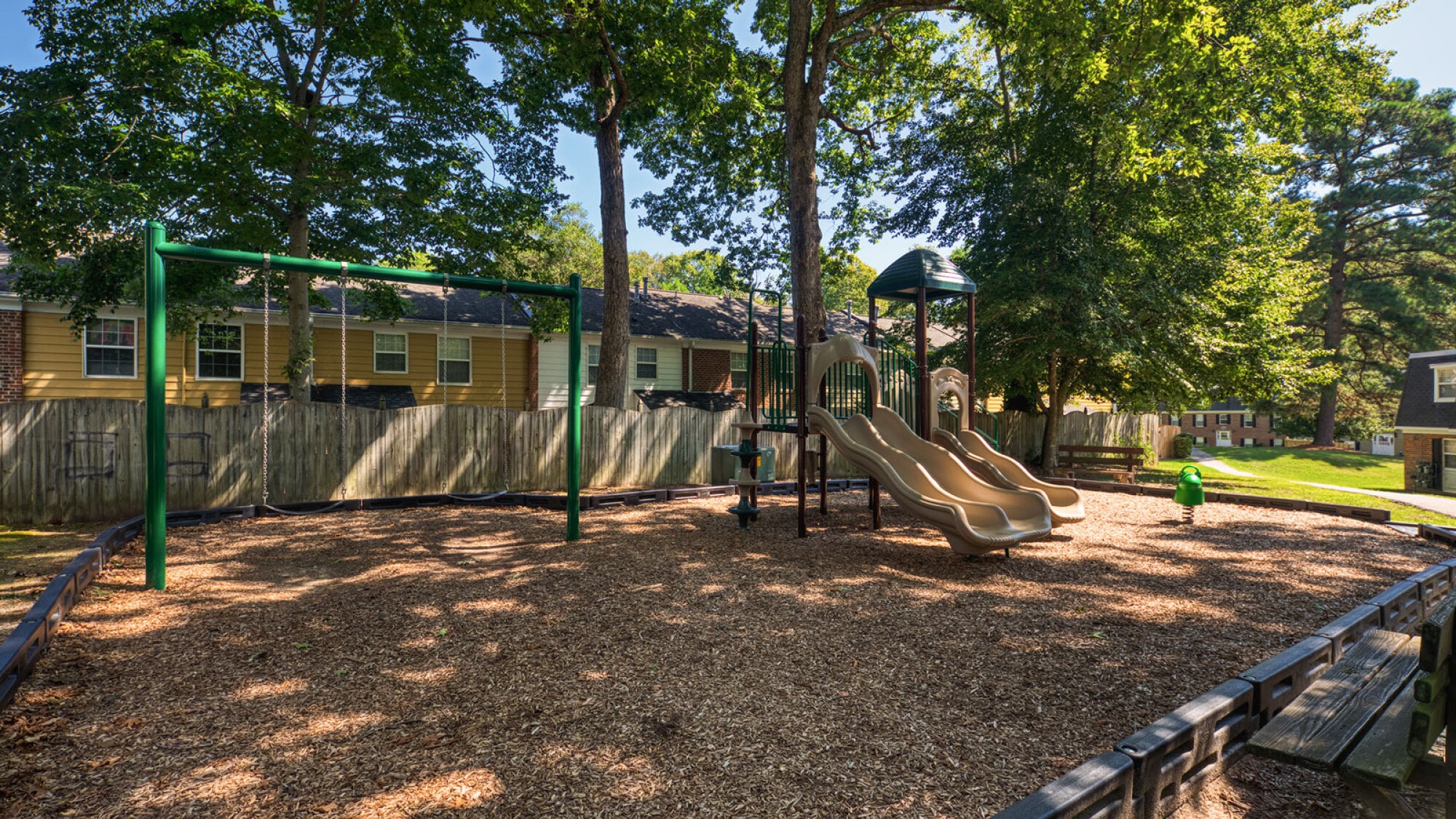 Outdoor playground with swings, two slides, and trees, surrounded by a wooden fence and houses in the background.