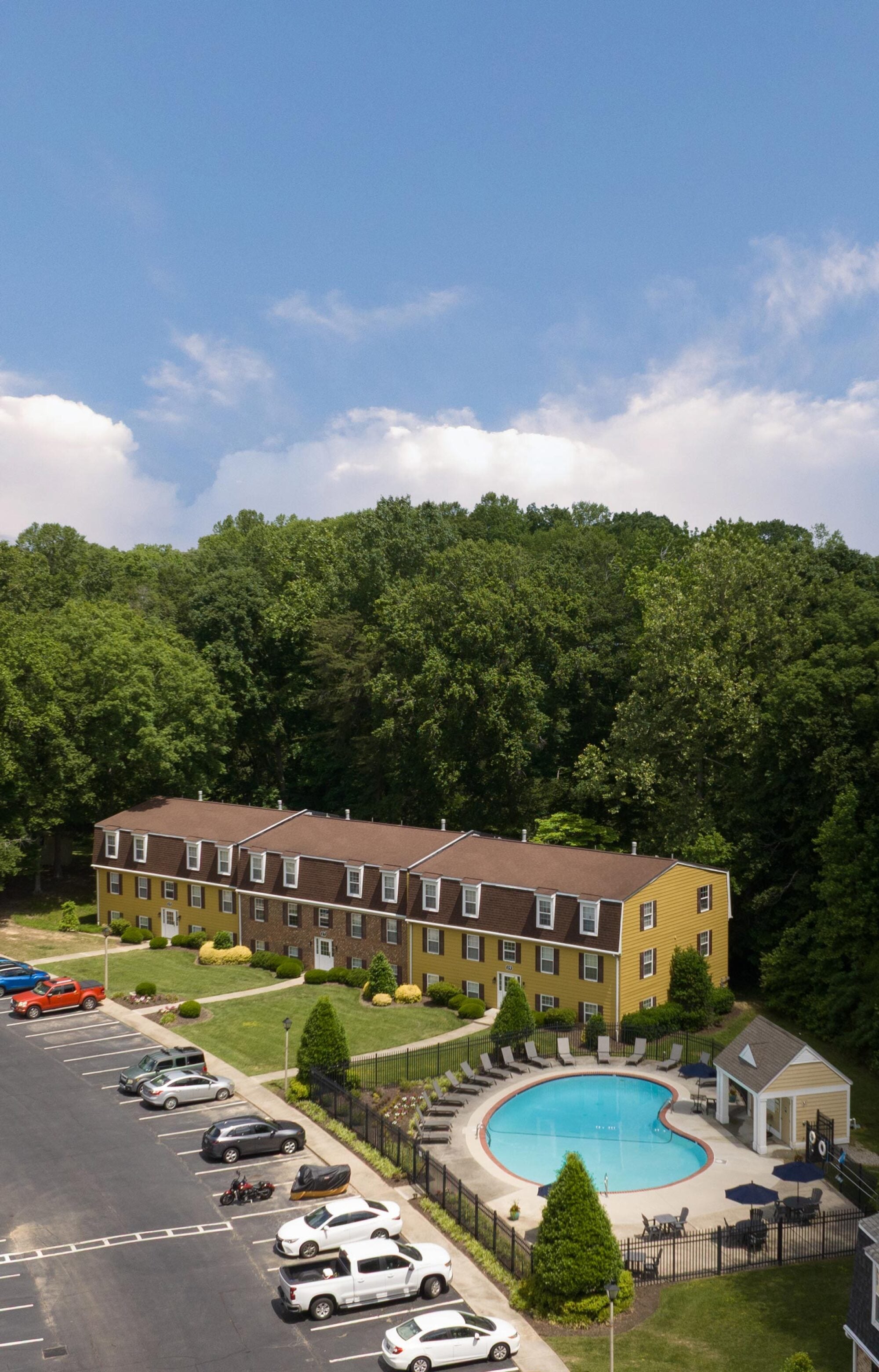 Aerial view of a yellow apartment building, parking lot, and round outdoor swimming pool surrounded by trees.