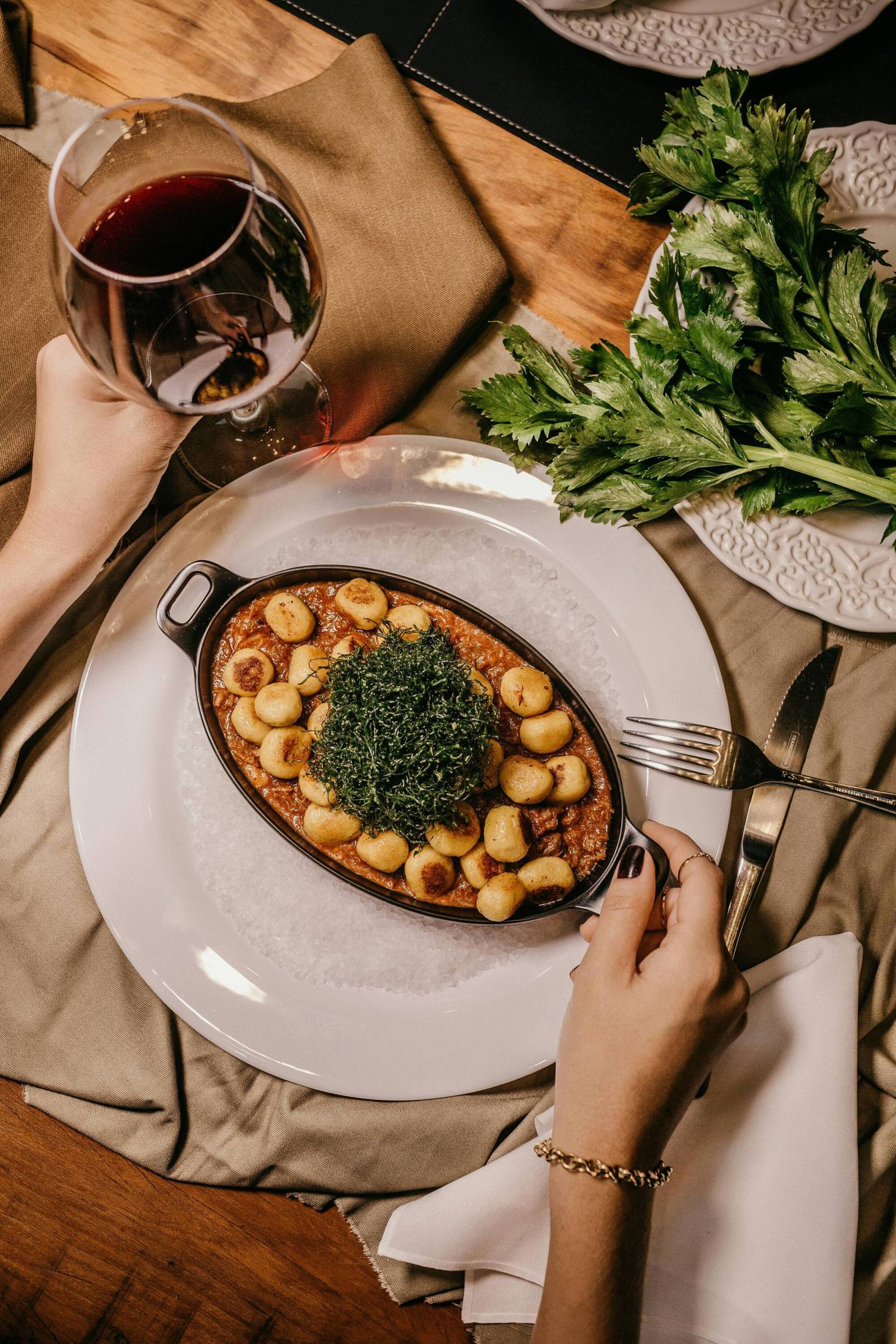 A person holds a glass of red wine beside a baked gnocchi dish with greens, set on a table with fresh parsley.