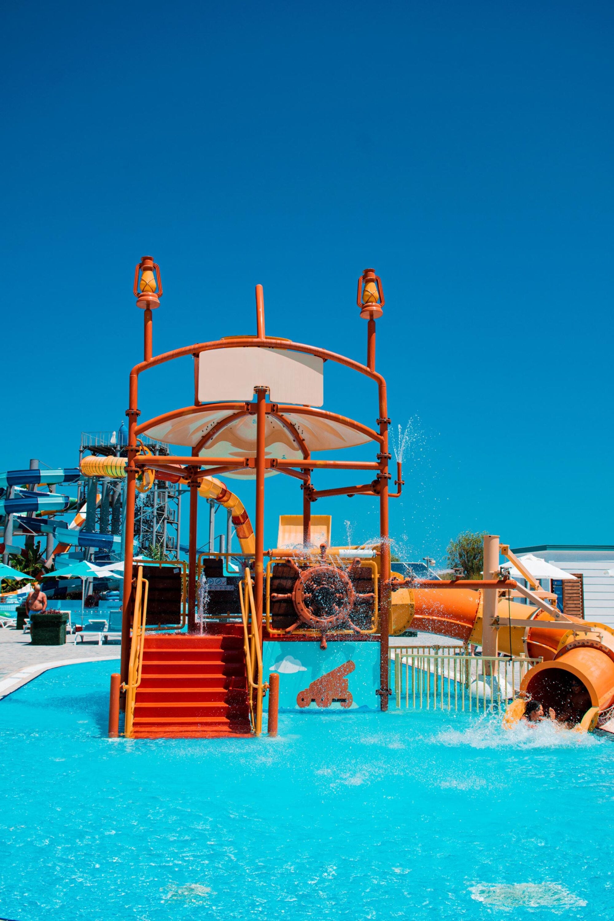 Bright orange water slide structure with dumping bucket, stairs, and splashing water under a clear blue sky.