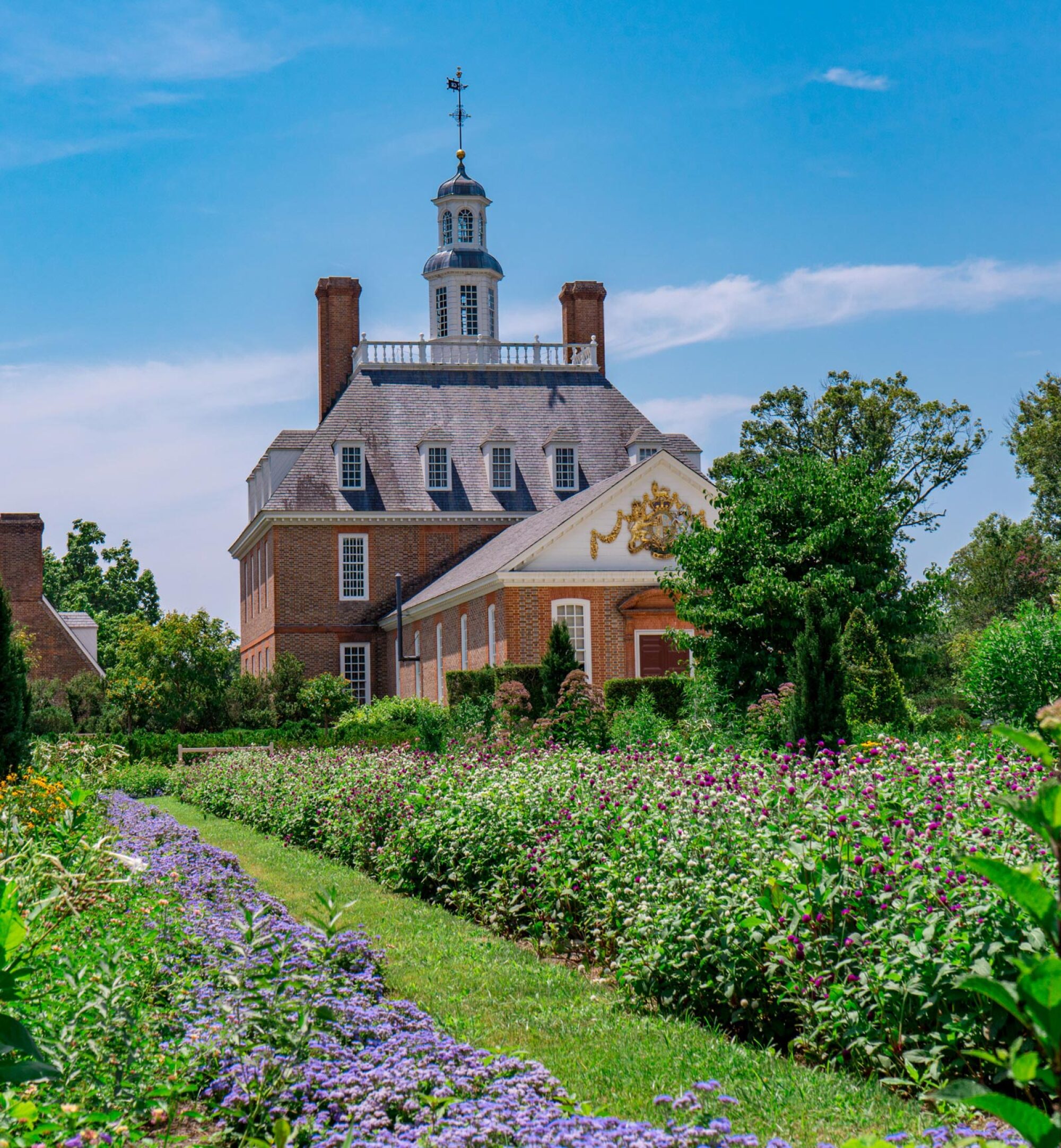 Brick colonial building with a cupola, surrounded by lush gardens and purple flowers under a blue sky.