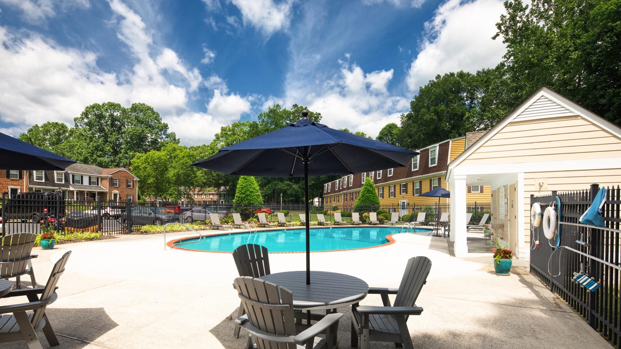 Outdoor pool area with lounge chairs, tables, and umbrellas, surrounded by trees and townhouse-style buildings.