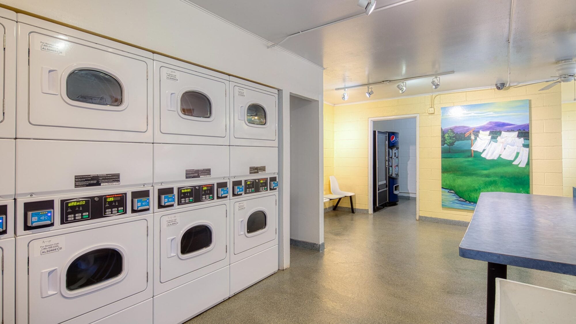 A clean laundry room with stacked washers and dryers, a folding table, chairs, and a colorful mural of clothes drying outside.