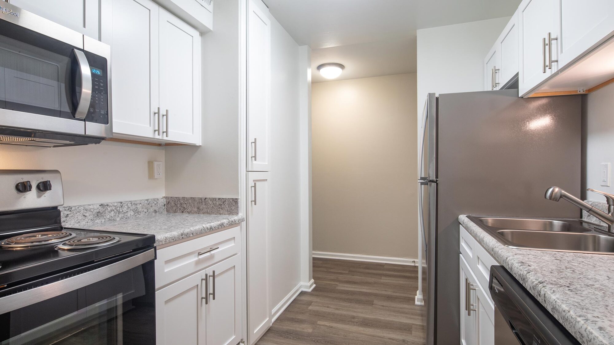 Modern galley kitchen with white cabinets, stainless steel appliances, and wood-look flooring under bright lighting.