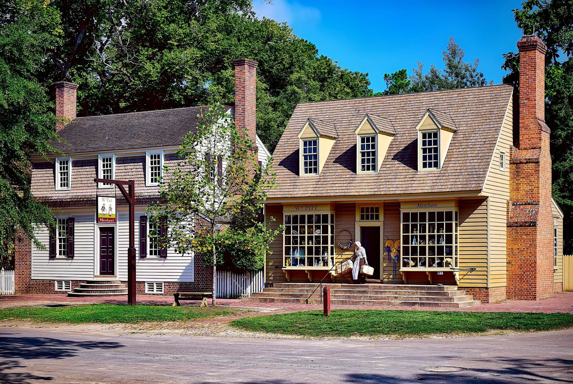 Two colonial-style buildings with brick chimneys, large windows, and a person sitting on the front steps under a bright blue sky.