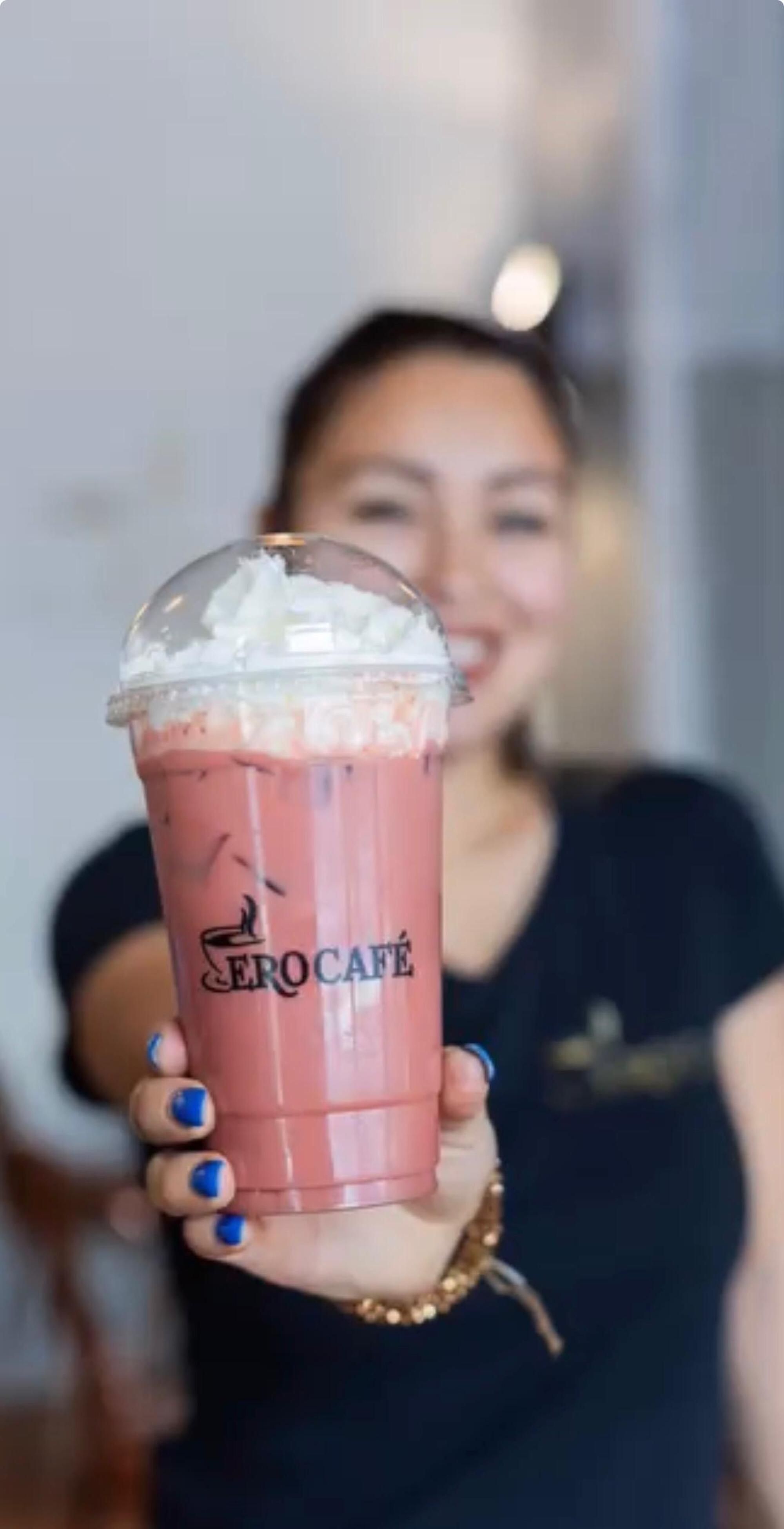 A woman holds out a pink iced drink with whipped cream in a clear cup labeled “Zero Café.”.