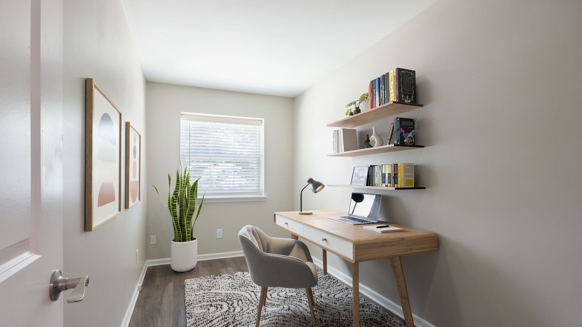 Minimalist home office with desk, chair, lamp, laptop, two shelves of books, a plant, and abstract art on the wall.