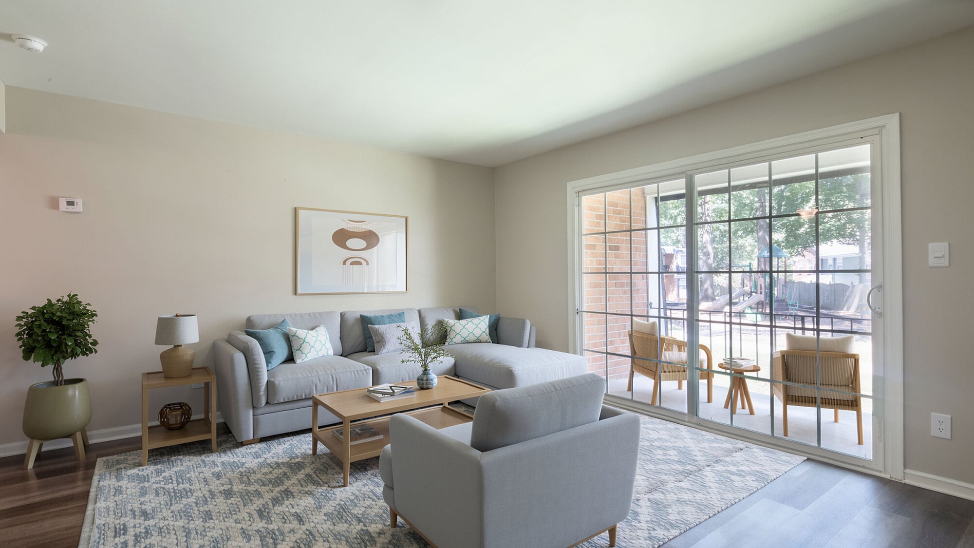 Modern living room with light gray furniture, abstract wall art, and a sliding glass door leading to a sunroom with chairs.