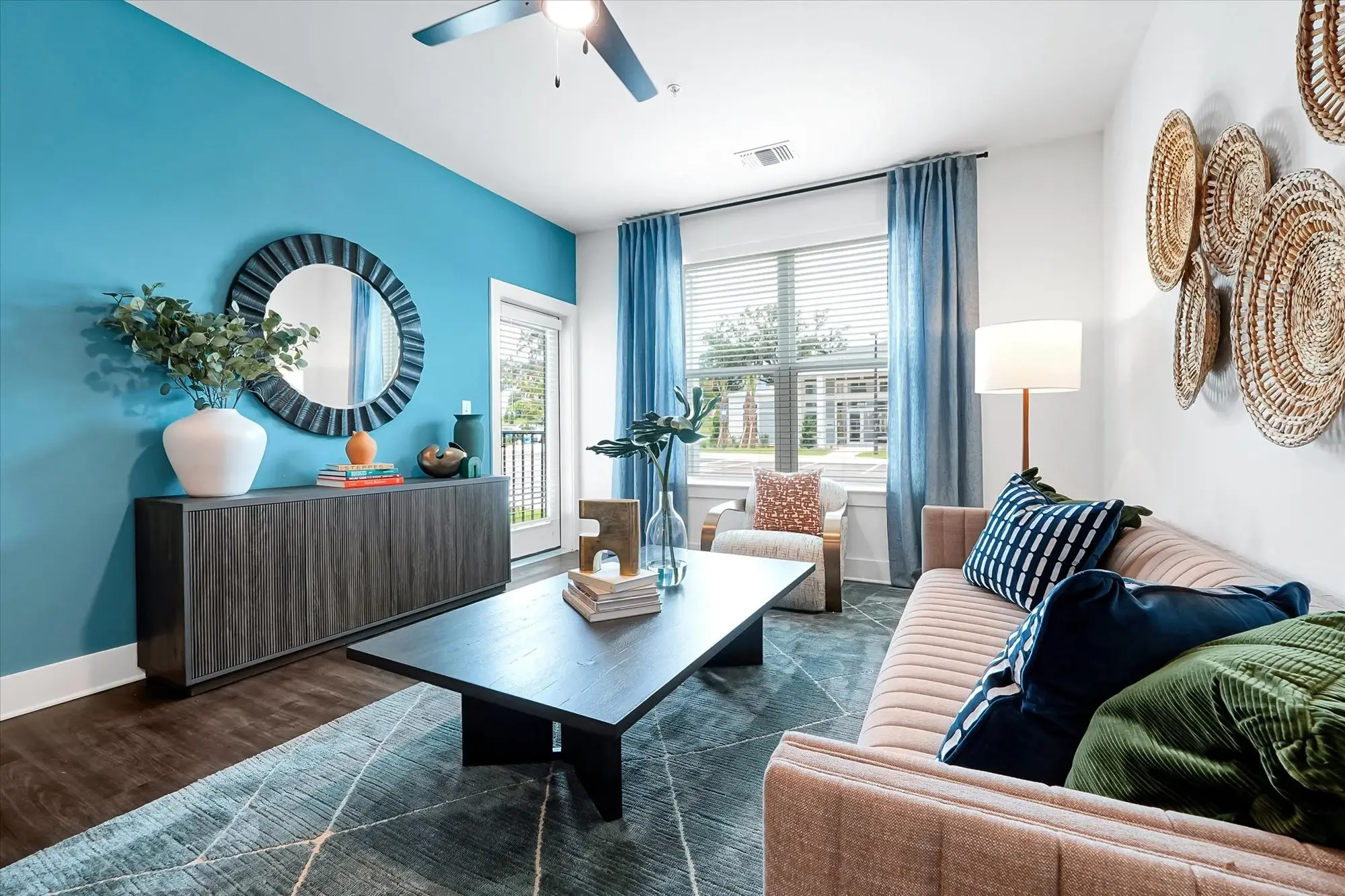 Modern living room with a blue accent wall, tan sofa, coffee table, and large window with blue curtains letting in natural light.