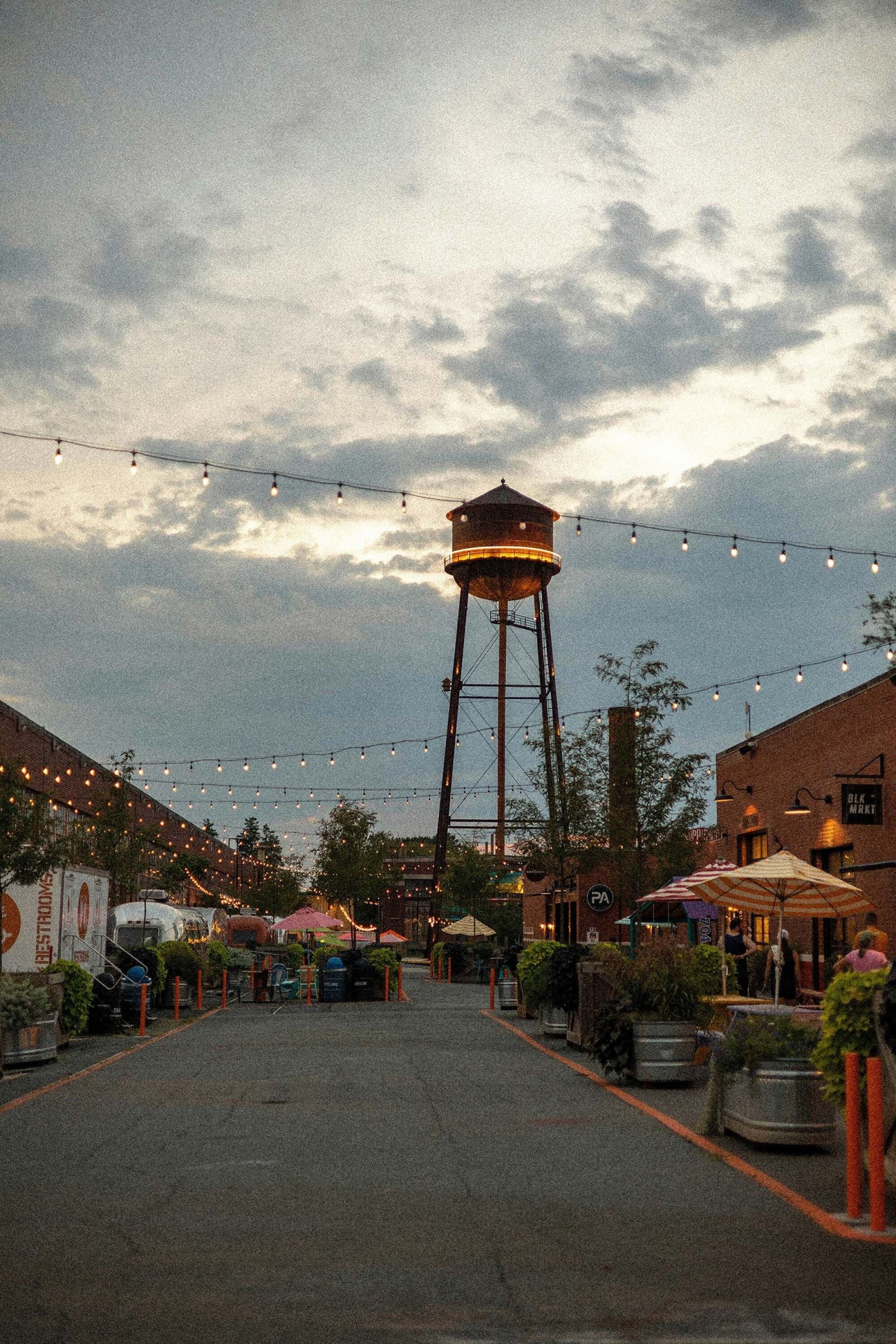 String lights over a brick market district with food trucks and patio umbrellas at dusk near Maple Run in Charlotte, NC.