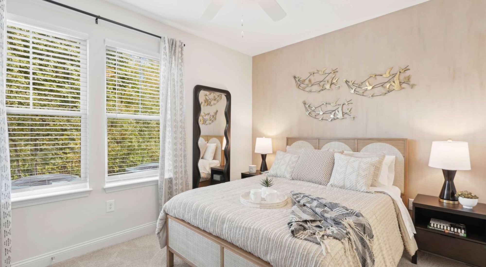 Bright bedroom with a bed, two bedside tables, wall art, a mirror, and windows with curtains letting in natural light.
