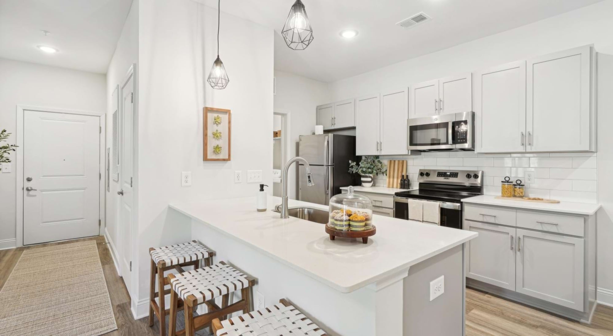 Modern white kitchen with gray cabinets, pendant lights, stainless steel appliances, and a breakfast bar with two woven stools.