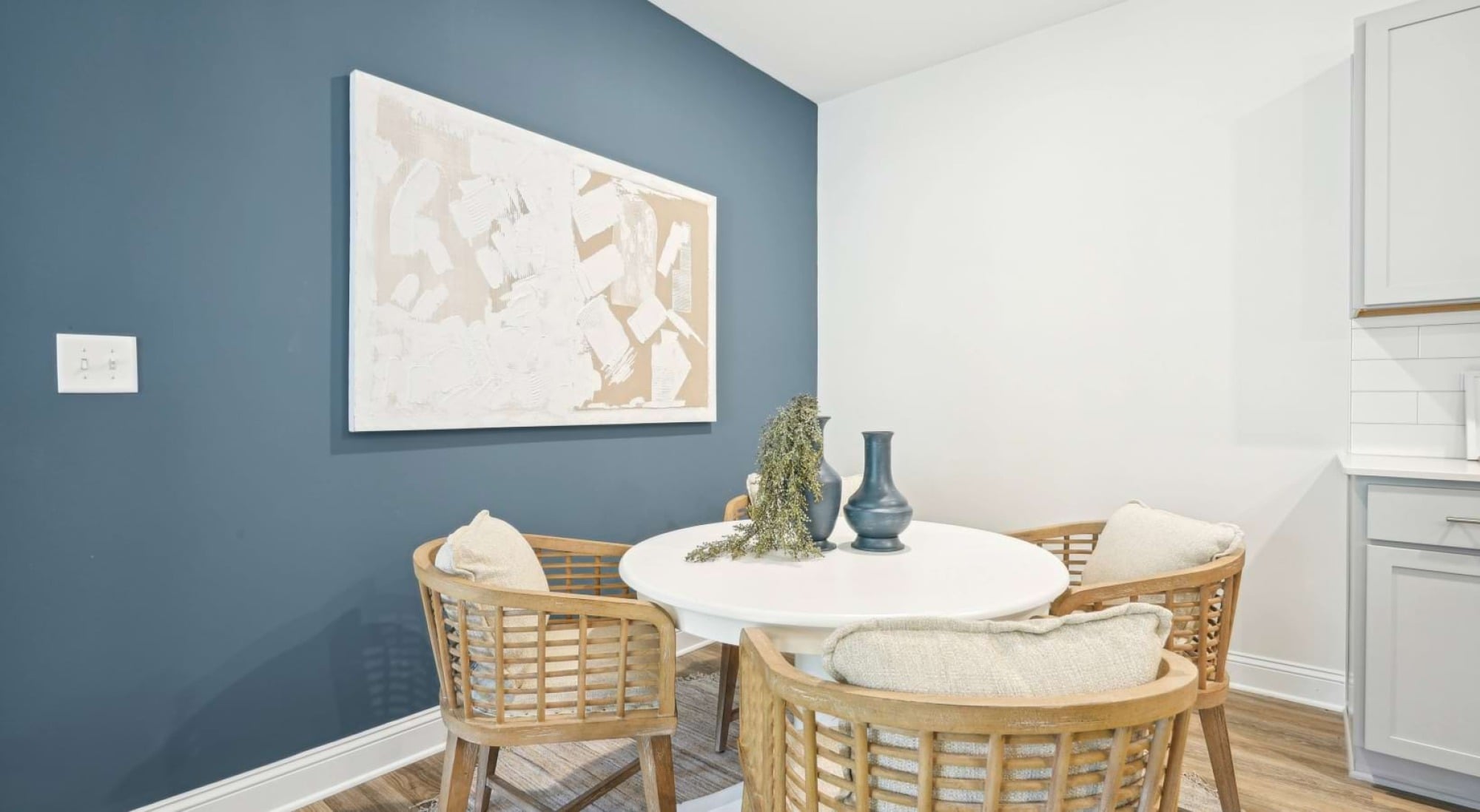 A round white table with four wicker chairs, a vase, and abstract art on a blue accent wall in a modern dining area.