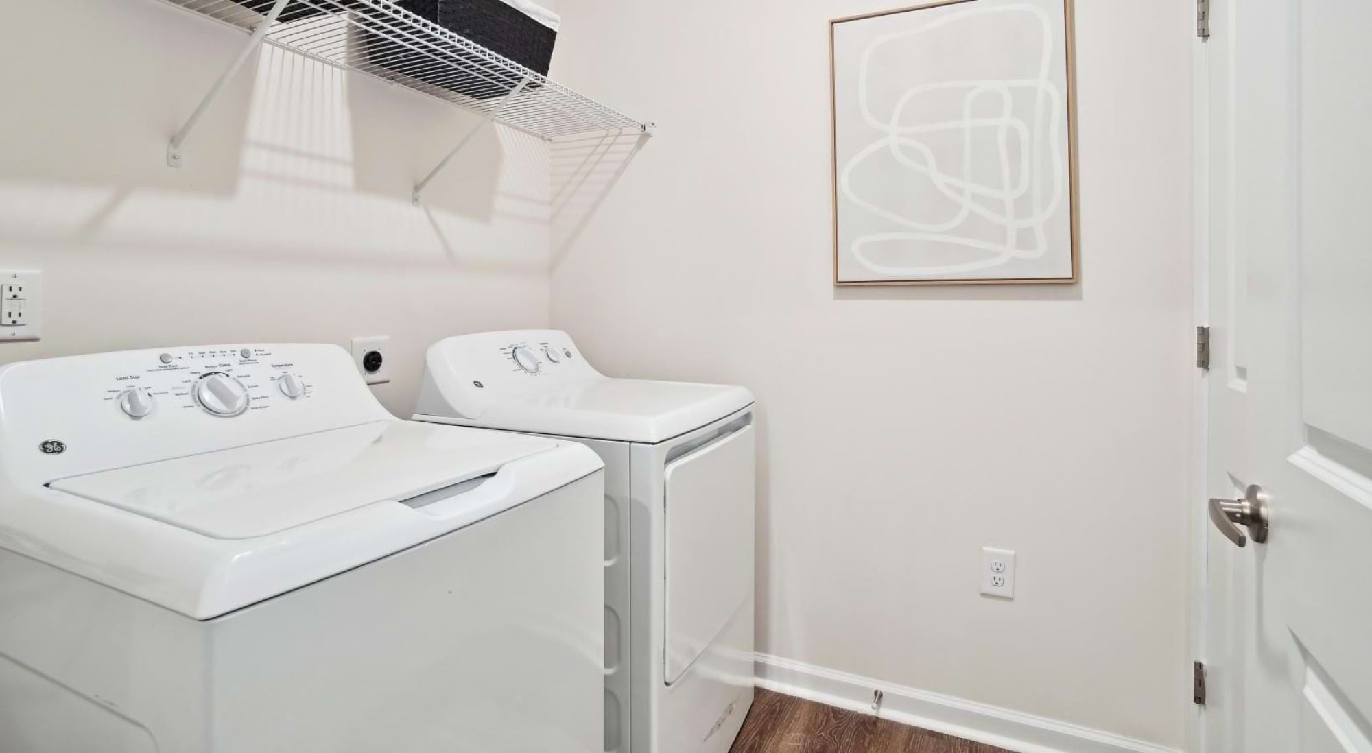 A laundry room with a washing machine, a dryer, wall art, and a wire shelf holding a black basket.