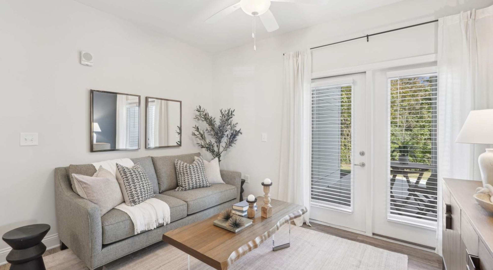 Bright living room with a gray sofa, coffee table, potted plant, and French doors opening to a balcony with greenery outside.