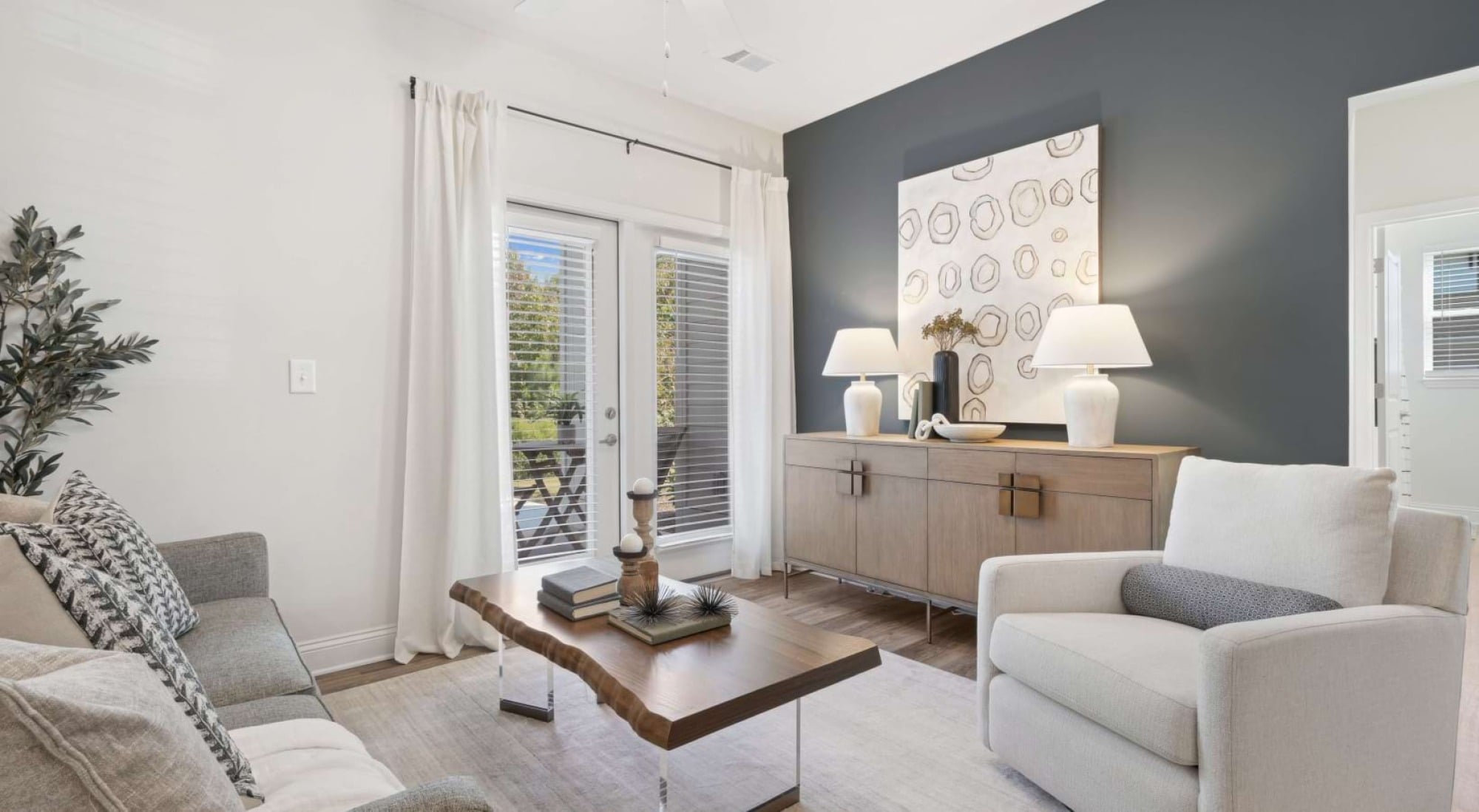Modern living room with gray accent wall, cozy seating, wood table, sideboard, lamps, art, and patio doors letting in sunlight.