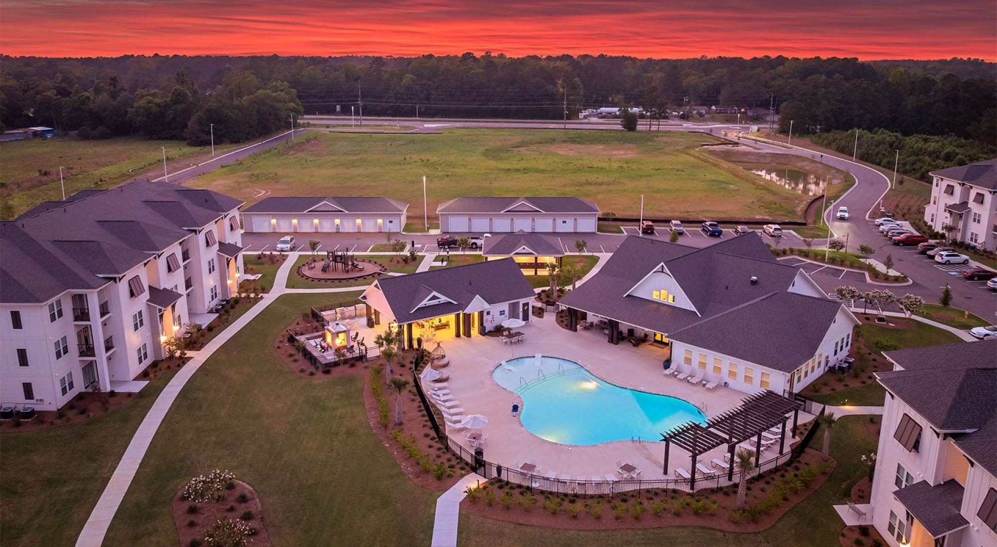 Aerial view of an apartment complex with a pool at sunset, surrounded by greenery and parking spaces.