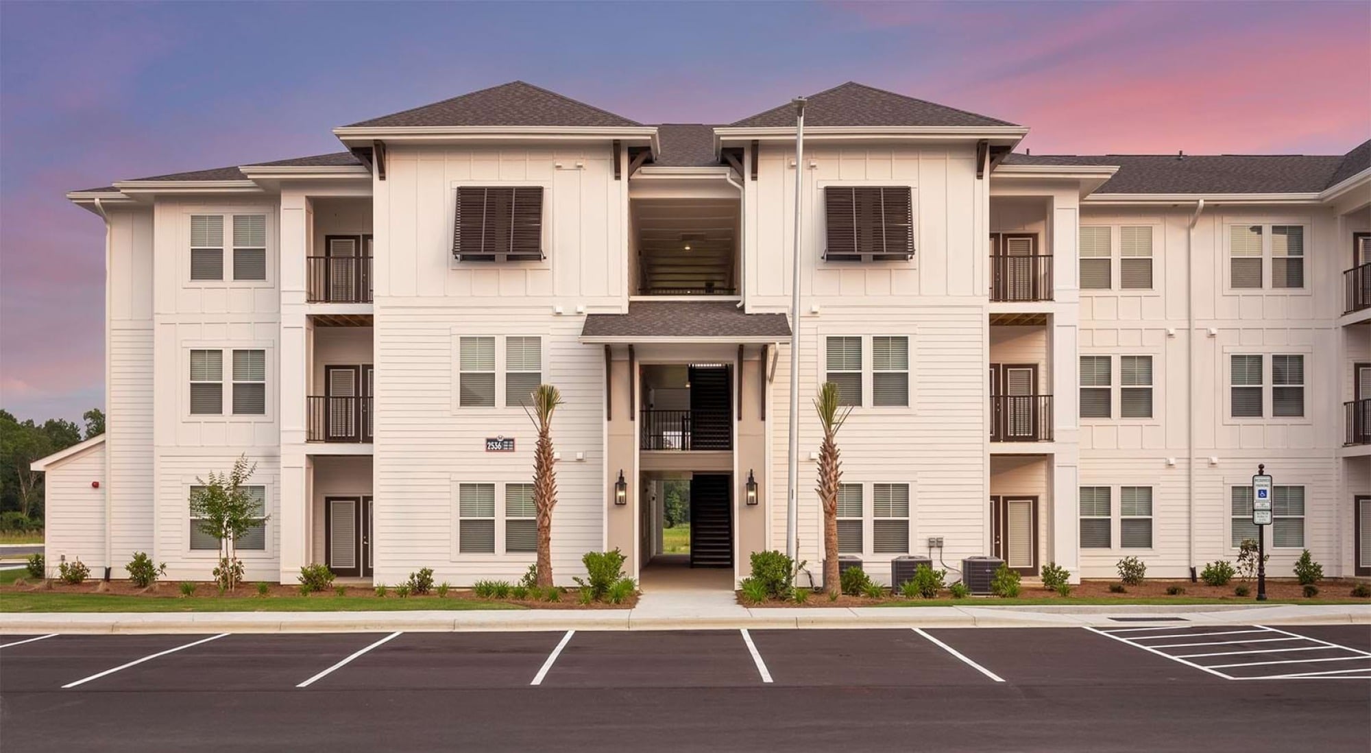 A modern three-story apartment building with white siding, black trim, balconies, and an empty parking lot at sunset.