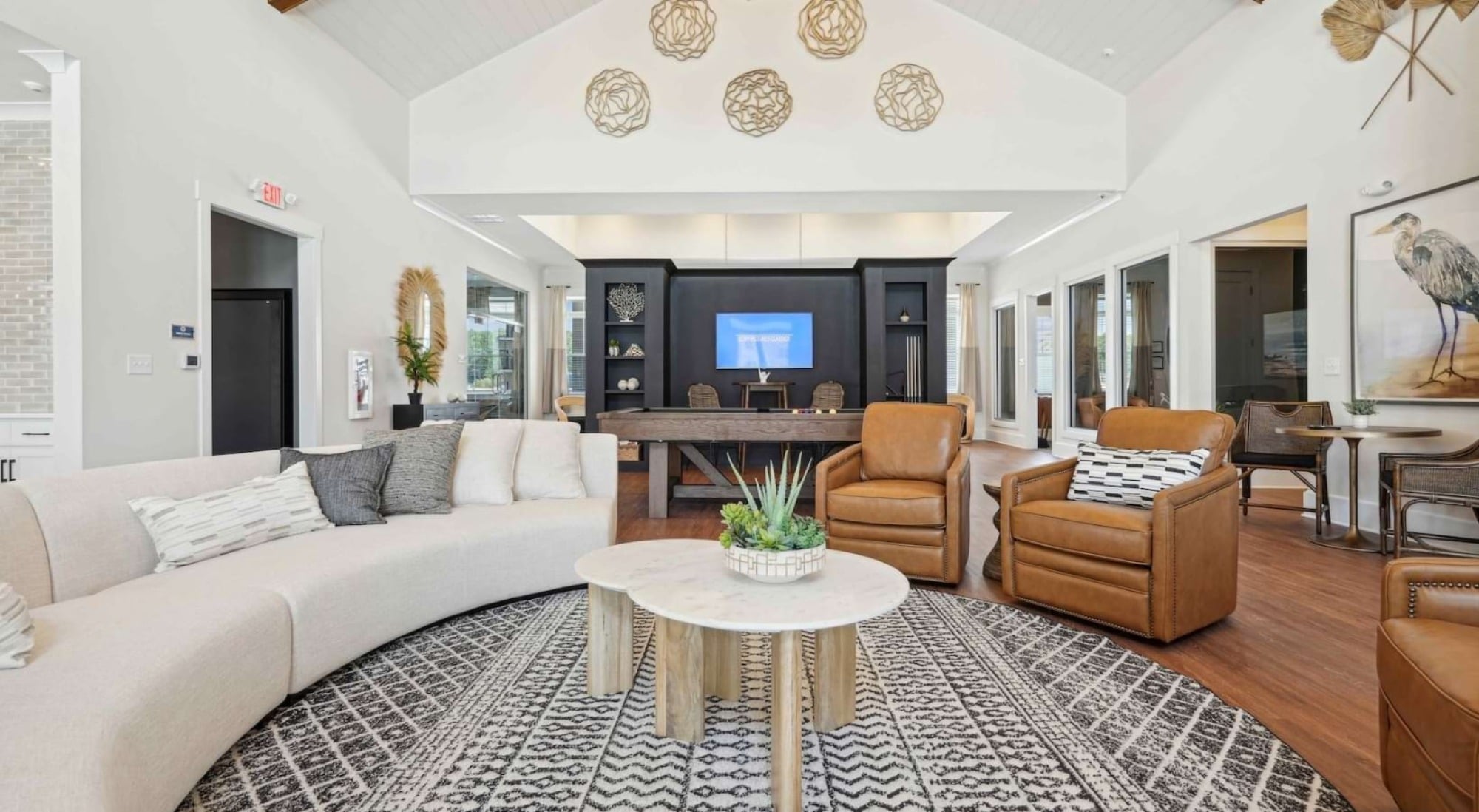 Modern lounge with a round sofa, tan armchairs, round coffee table, rug, and decorative lights hanging from vaulted ceiling.