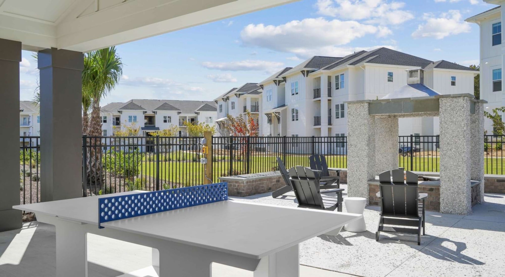 A white ping pong table on a patio with black chairs, overlooking modern white apartment buildings and a fenced green lawn.