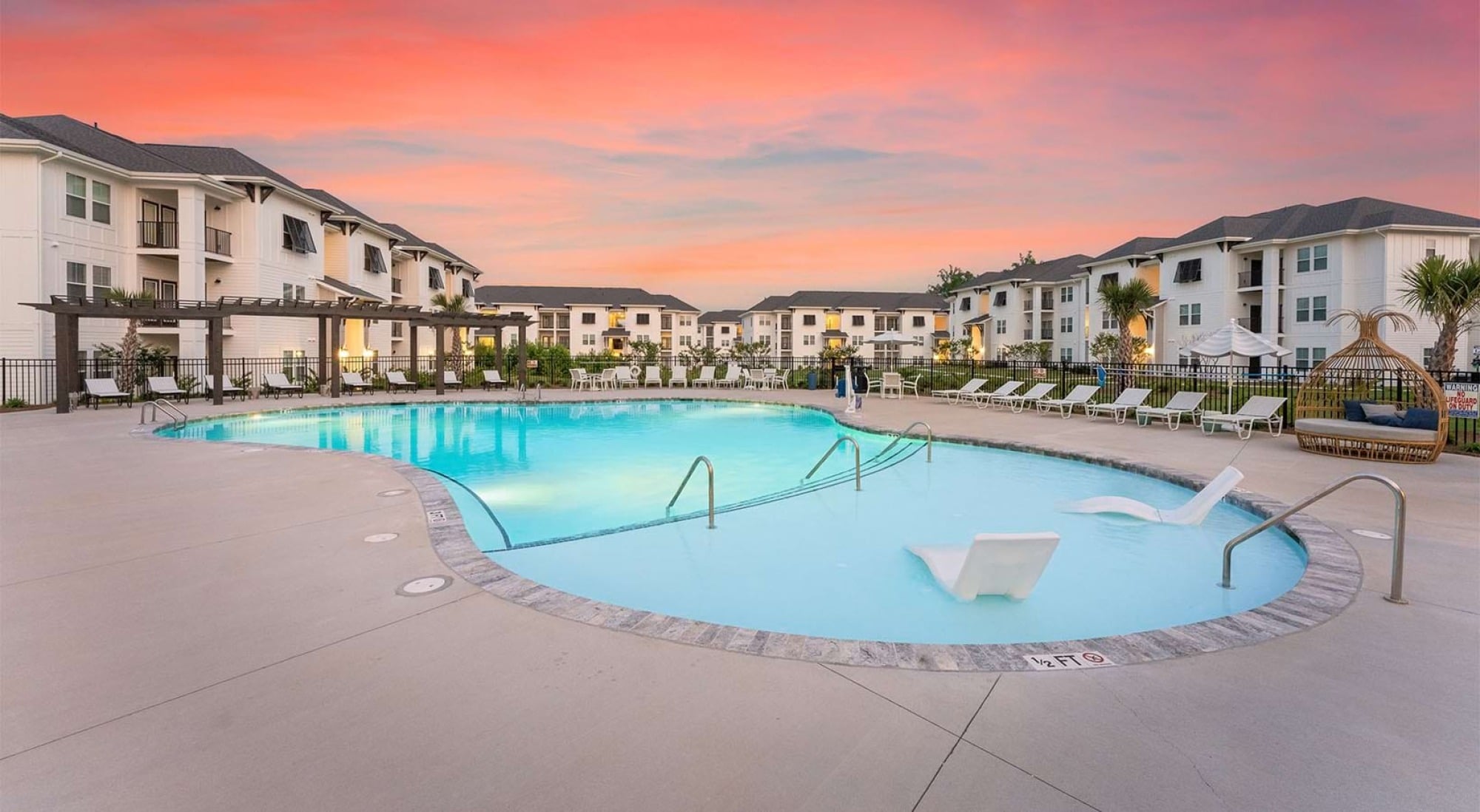 Swimming pool at an apartment complex with lounge chairs and palm trees under a pink sunset sky.