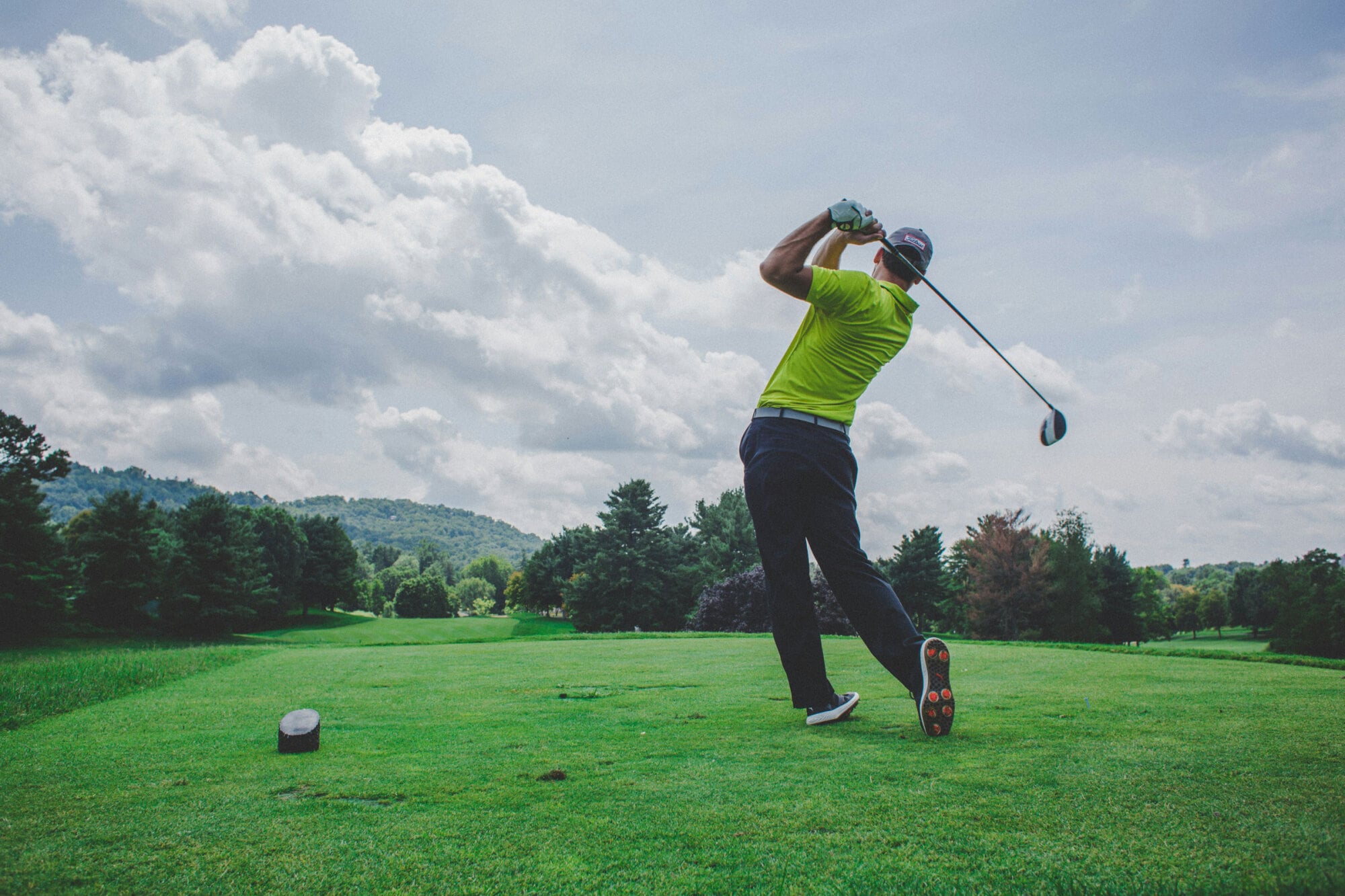 A golfer in a yellow shirt swings a golf club on a green course with trees and cloudy sky in the background.