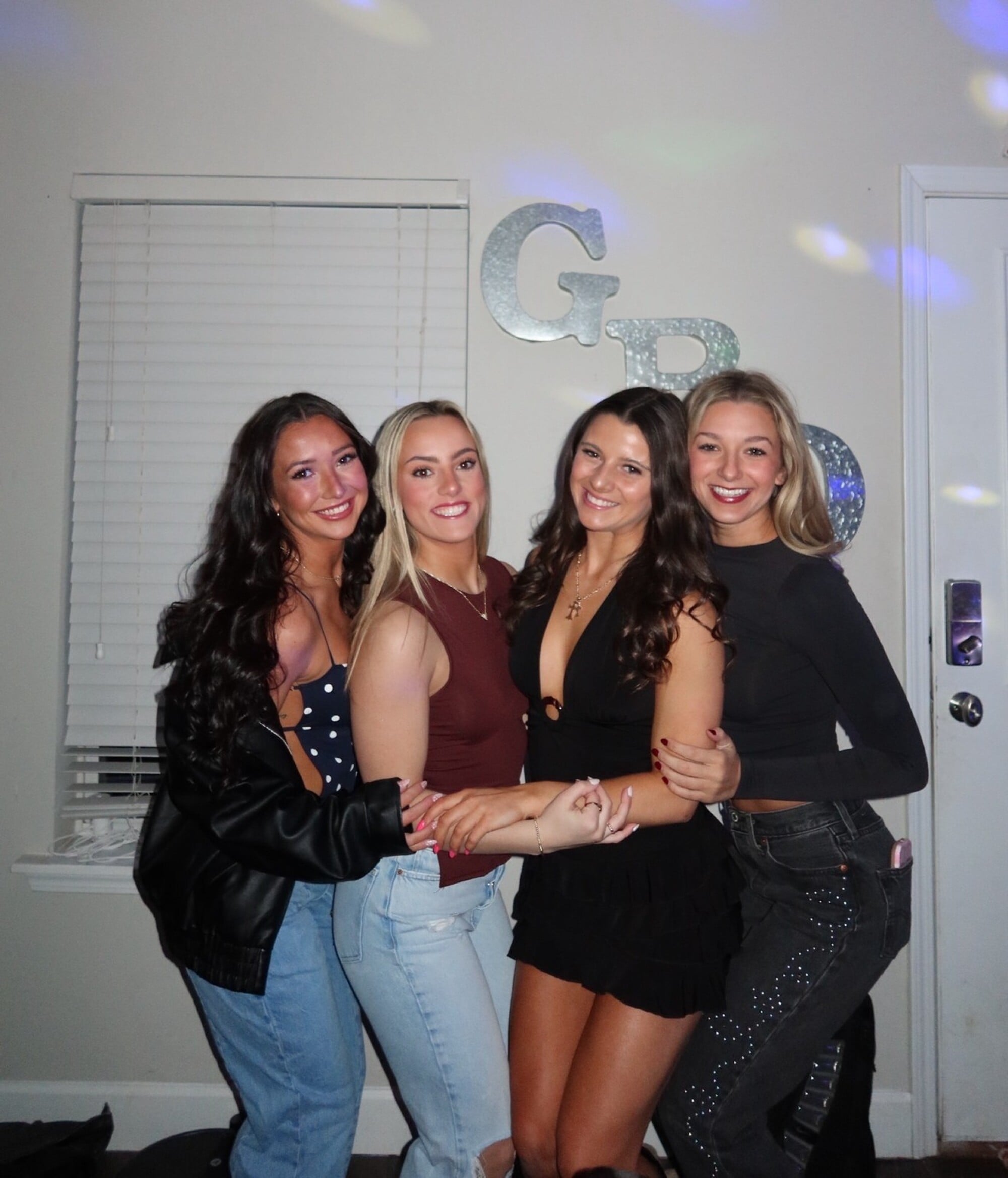 Four college friends smiling with their arms around each other at an evening get-together indoors.
