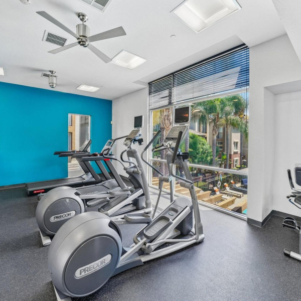 Cardio equipment at Ontario Town Square Townhomes in Ontario, CA, including ellipticals and a recumbent bike beside a sunlit window.