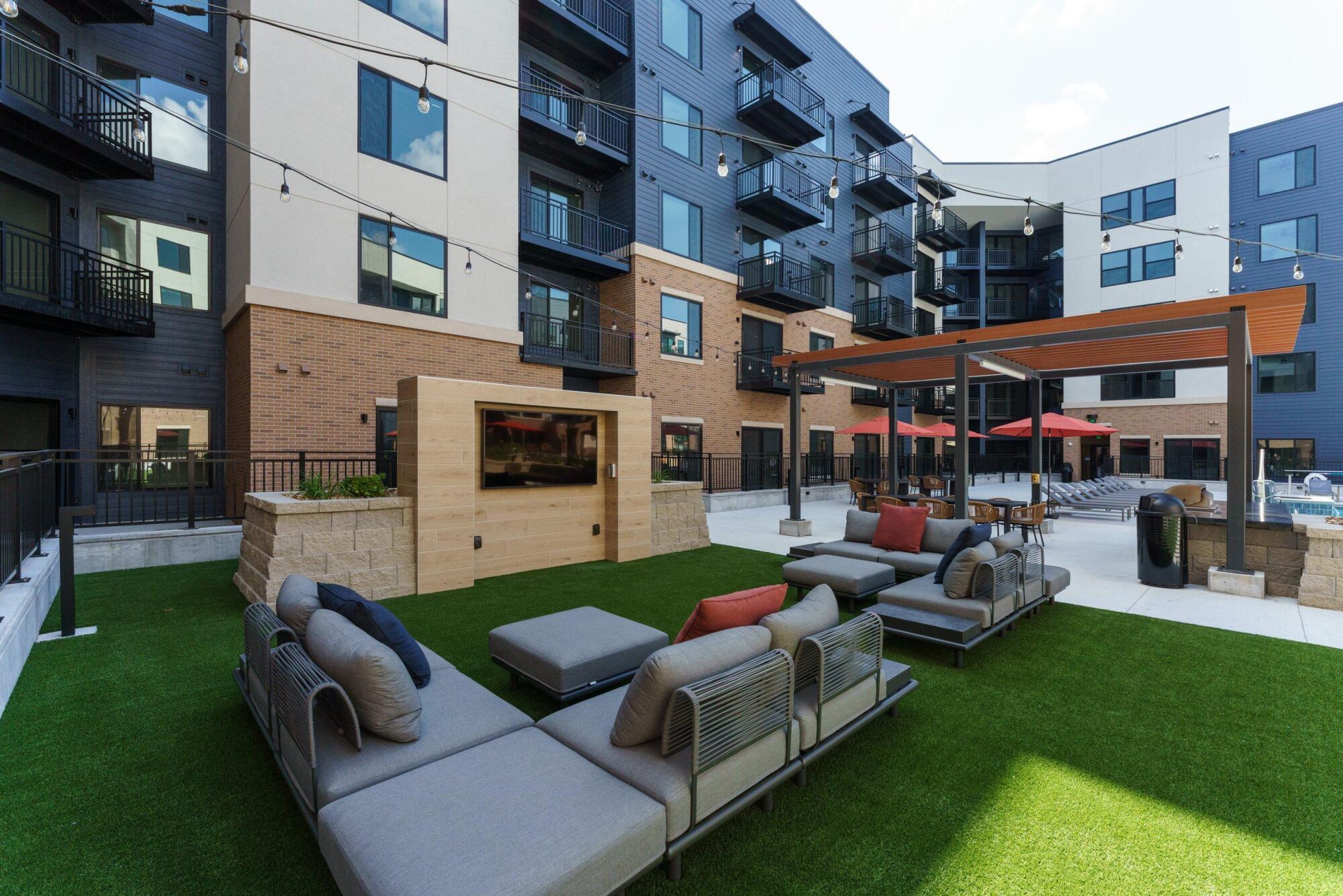 Courtyard lounge with outdoor sectional seating on turf, a stone TV wall, a pergola and string lights overhead