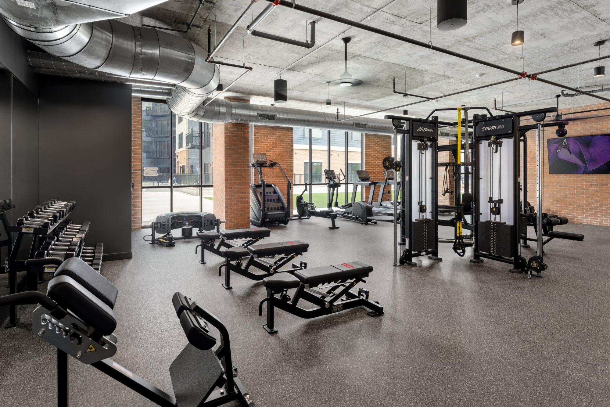 Fitness center with cable machines, adjustable benches, dumbbells and cardio equipment under exposed ductwork