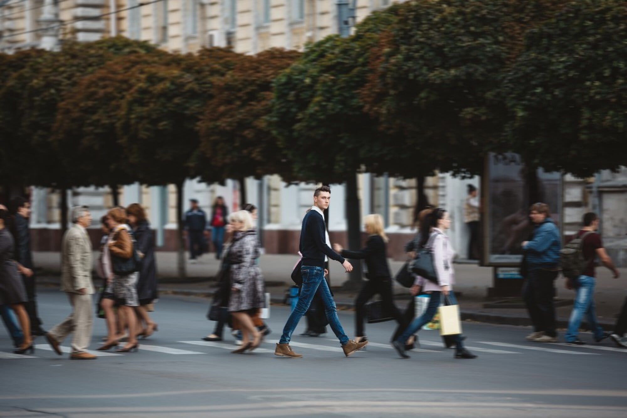 A man in blue jeans walks confidently while others cross a city street, blurred in motion. Large trees and buildings are in the background.